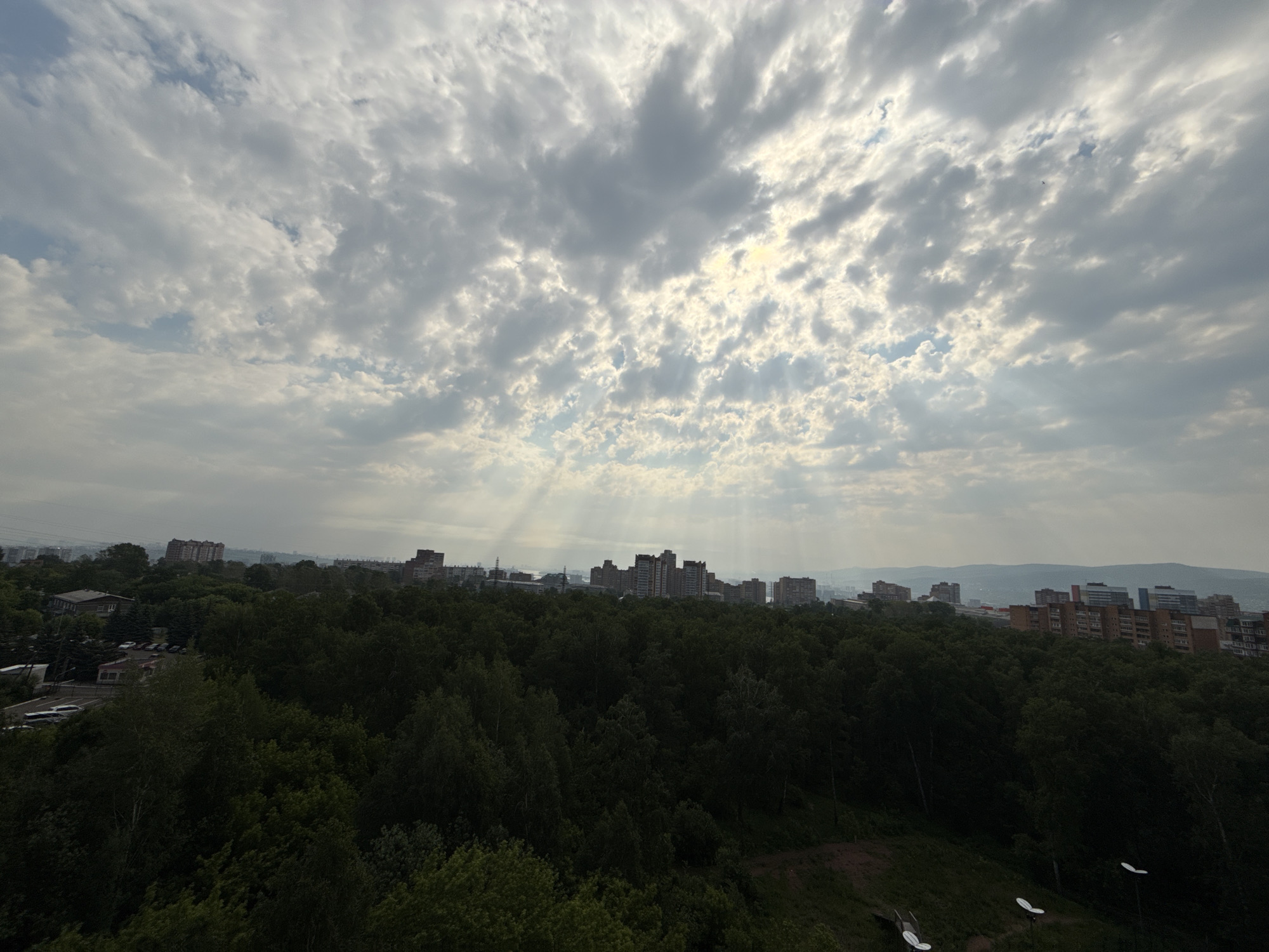 The image captures a panoramic view of a city skyline in Krasnoyarsk, Russia, dominated by a dense forest in the foreground and a cluster of apartment buildings in the middle ground. The sky is overcast with dramatic clouds, from which rays of sunlight pierce through, creating a diffused and atmospheric lighting effect. The city appears to be situated on rolling hills, with more distant hills fading into a hazy background. The foreground consists of a lush, dark green forest, suggesting a natural area within or bordering the urban landscape. The apartment buildings are of varying heights and architectural styles, typical of Soviet-era and post-Soviet construction, with some exhibiting a reddish-brown brick facade. A few cars are visible in a parking area near one of the buildings, indicating some level of human activity. The overall impression is one of a city integrated with nature, under a dynamic and somewhat subdued sky.