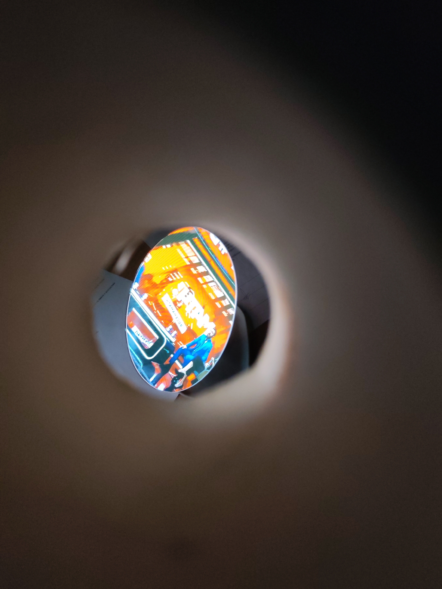 The image shows a blurred view through a hole, possibly a peephole or a narrow opening. The scene visible through the hole is a brightly colored image, seemingly a depiction of a person in a blue outfit with orange and white elements in the background. It's difficult to discern details about the location or precise subject matter of the image within the hole due to the limited view and distortion. The surroundings of the hole are out of focus and appear to be a neutral color, giving no contextual information about its location or the immediate environment. Without further information or a clearer view, the scene's connection to the city of Shcheleiki, Russia is indeterminable.