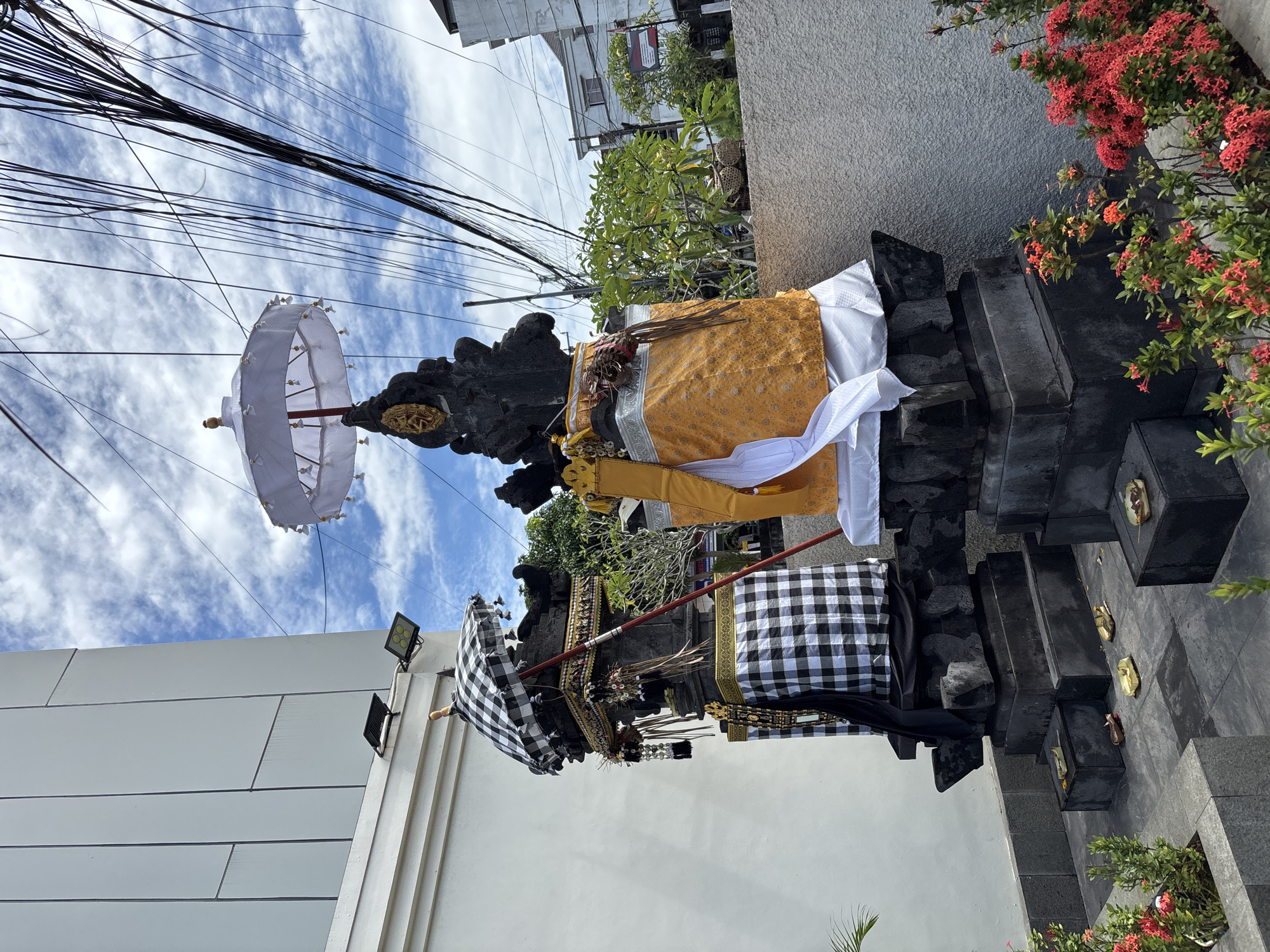 A stone shrine in Kuta Selatan, Indonesia, is adorned with offerings and fabrics. The shrine itself is constructed of dark gray stone and consists of several stepped levels. It is decorated with a yellow patterned cloth, white cloth, and a black and white checkered cloth. In front of the shrine, at the base of the stepped levels, are scattered small offerings of what appears to be fruit and flowers. Two ceremonial umbrellas are present, one white and the other black and white checkered, adding to the shrine's ornate presentation.
The background includes a building with a white wall, a gray concrete wall, and overhead power lines against a blue sky with scattered clouds. Red flowering plants are to the right of the shrine.
