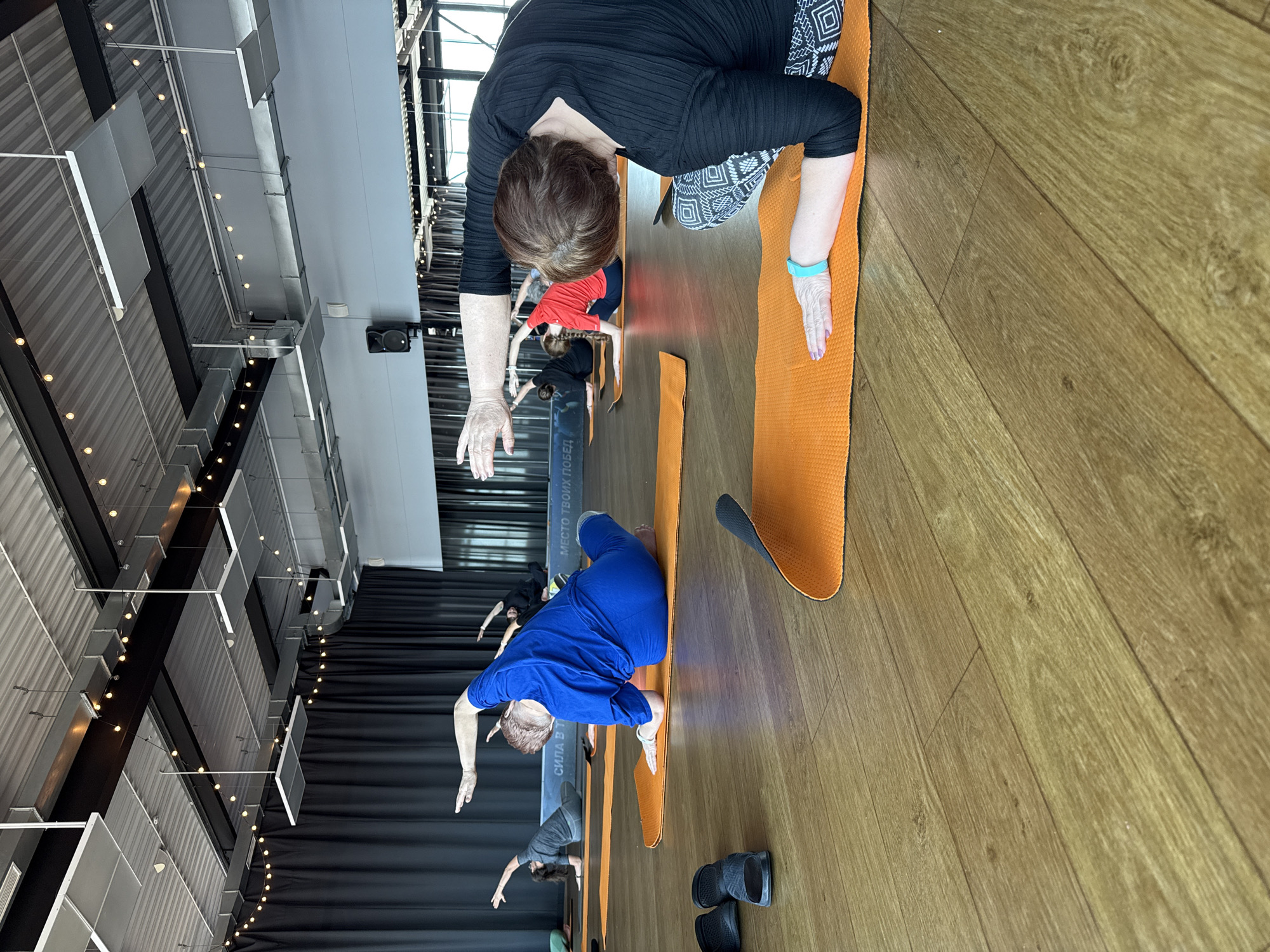 The image shows a yoga class in progress in Votkinsk, Russia. Multiple people are on orange yoga mats performing a bending exercise. They are lined up in a single row, with a woman in a black top and patterned pants in the foreground, followed by other participants dressed in blue, red, and gray attire. The setting appears to be an indoor studio with wooden floors. Above, there is a high ceiling with visible industrial-style lighting fixtures and black drapes covering the windows. A few pairs of shoes are scattered near the yoga mats. Text is visible on one of the yoga mats.