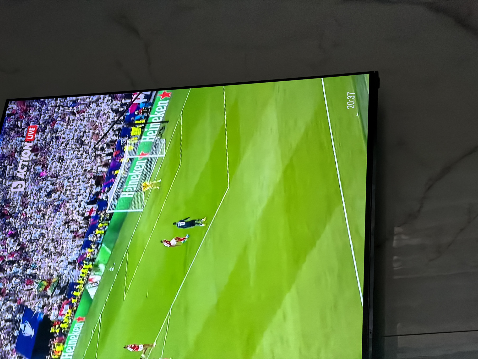 The image shows a television screen displaying a live soccer game. The camera focuses on a field with two players near midfield. A goalkeeper is positioned near the net. The stands are packed with spectators. Advertisements for "Heineken" are visible along the sidelines. The game time on the screen reads "20:37". The photo was taken in a private residence in Asenovgrad, Bulgaria.