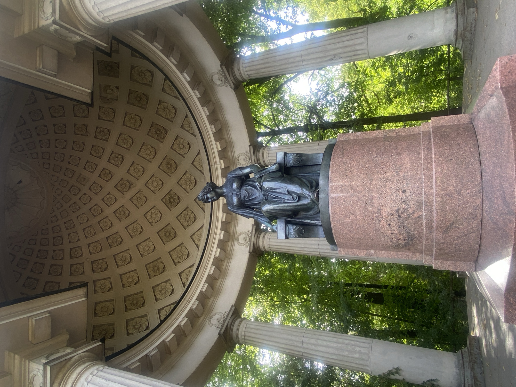 A full shot shows a bronze statue of a seated woman in a classical dress and crown, situated on a granite pedestal. The statue is positioned within a circular colonnade that has fluted columns supporting a domed ceiling decorated with a repeating floral pattern. The colonnade is surrounded by lush green trees, with sunlight filtering through the leaves. The location is identified as the monument to Empress Maria Feodorovna in the Pavlovsk Park, Russia.