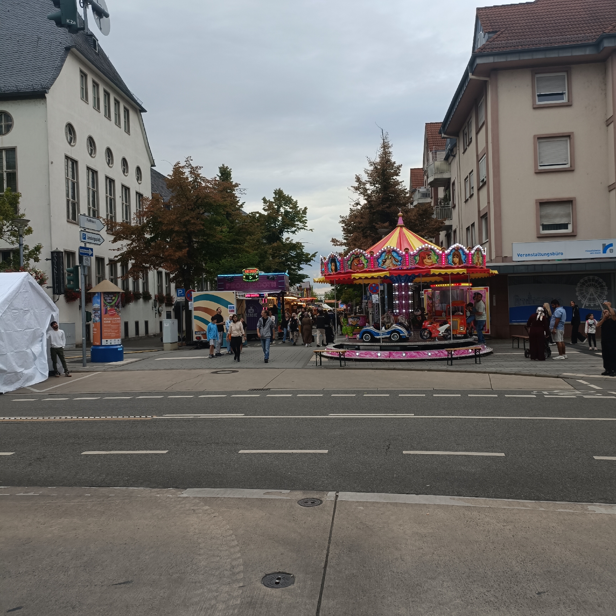 The image depicts a street scene in Rüsselsheim am Main, Germany, on a cloudy day. In the foreground, a street with multiple lanes and white markings is visible. Beyond the street, a pedestrian walkway is lined with buildings and a festive atmosphere. A colorful carousel, a "Photo Booth" truck, and other stalls suggest a fair or event is taking place. Several people are walking along the walkway, some observing the attractions, while others are engaged in conversations. A white tent is set up on the left side of the walkway, and a man is standing near it. The buildings in the background vary in architectural style, with some appearing older and others more modern. Street signs indicating "Rathaus" (City Hall) and "Landingsplatz" (Landing Place) are visible on the left, providing context for the location. The sky is overcast, casting a diffused light over the scene.