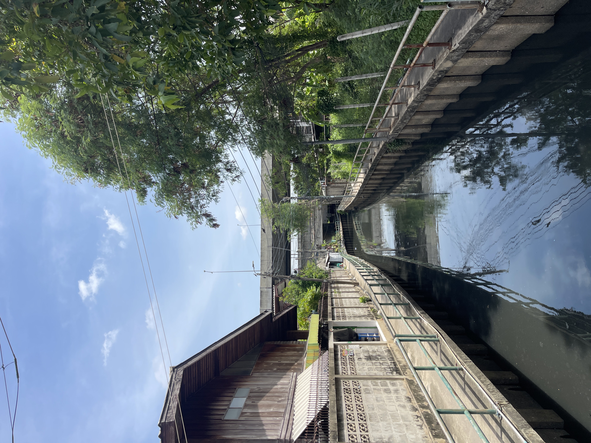 The image depicts a canal in Bangkok, Thailand. On the left side of the canal, there is a weathered wooden building with a sloping roof and a striped awning. A concrete wall with a decorative pattern runs along the edge of the building. A paved walkway with a green metal railing follows the canal. On the right side of the canal, there is dense green foliage and trees, with some electrical wires running overhead. The canal water is dark and reflects the sky and surrounding trees. In the distance, an elevated highway or bridge structure is visible over the canal. The sky is blue with scattered white clouds. The scene appears to be a residential or commercial area alongside a waterway in a tropical urban environment. There are no people or animals visible in the image.