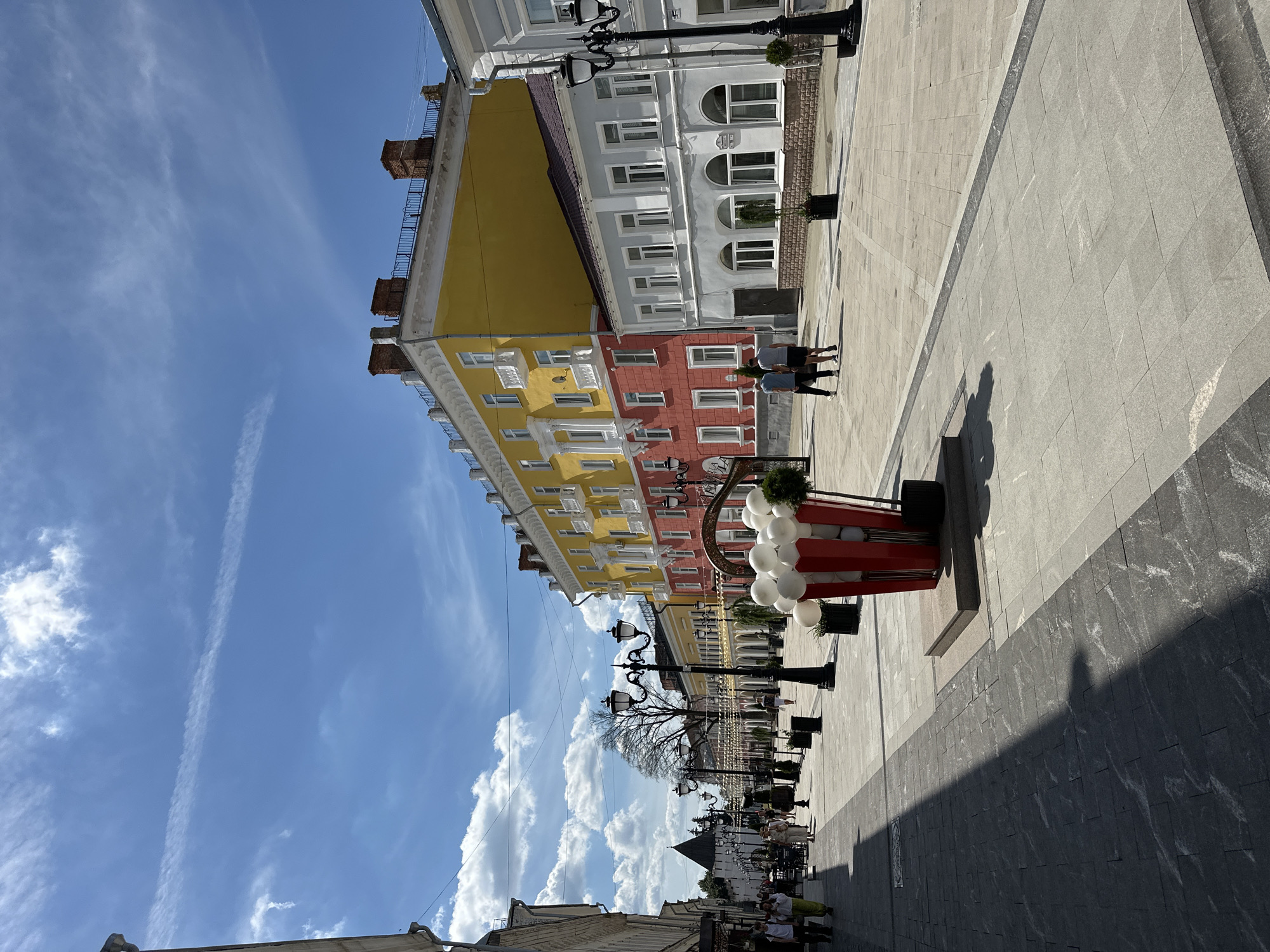 The image depicts a sunny day on a wide, pedestrianized street in a city. The street is paved with light-colored rectangular tiles, with darker gray paving stones forming a border and accentuating sections of the walkway. Shadows from buildings and trees stretch across the pavement.
On the right side of the street, a multi-story building features a bright yellow upper section and a salmon-pink lower section, with white balconies and architectural detailing. Adjacent to it is a lighter blue-gray building with arched windows. Ornate streetlights with multiple lamps line the walkway. In the foreground on the right, a large decorative element resembling a red and white popcorn bucket is filled with white balloons.
On the left side of the street, buildings are visible, and further down the street, a medieval-style tower with dark stone walls stands against the blue sky. The street is lined with trees and more decorative lights strung overhead. Several people are walking along the street, some in small groups, dressed in casual summer clothing. Two individuals are walking away from the camera in the middle ground on the right. The sky is a bright blue with scattered white, wispy clouds.