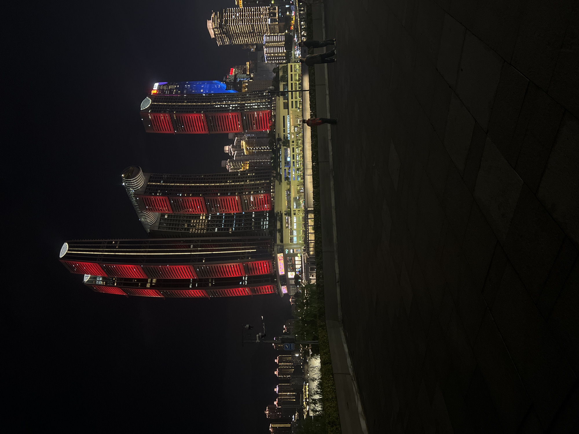 In the city of Jiangbeicheng Street, China, at night, several illuminated skyscrapers pierce the dark sky. The prominent structures are tall, modern buildings with red and white lighting accentuating their facades. One building features a unique circular top with lights. To the right, another skyscraper glows with blue and white lights. In the middle ground, a lower, multi-story building complex is visible, also lit up with yellow and white lights. In front of this complex, several people stand on a paved area. A few smaller buildings with illuminated windows are visible in the far distance on the left, suggesting a waterfront or riverfront setting. The foreground consists of a dark, paved area with visible square patterns.