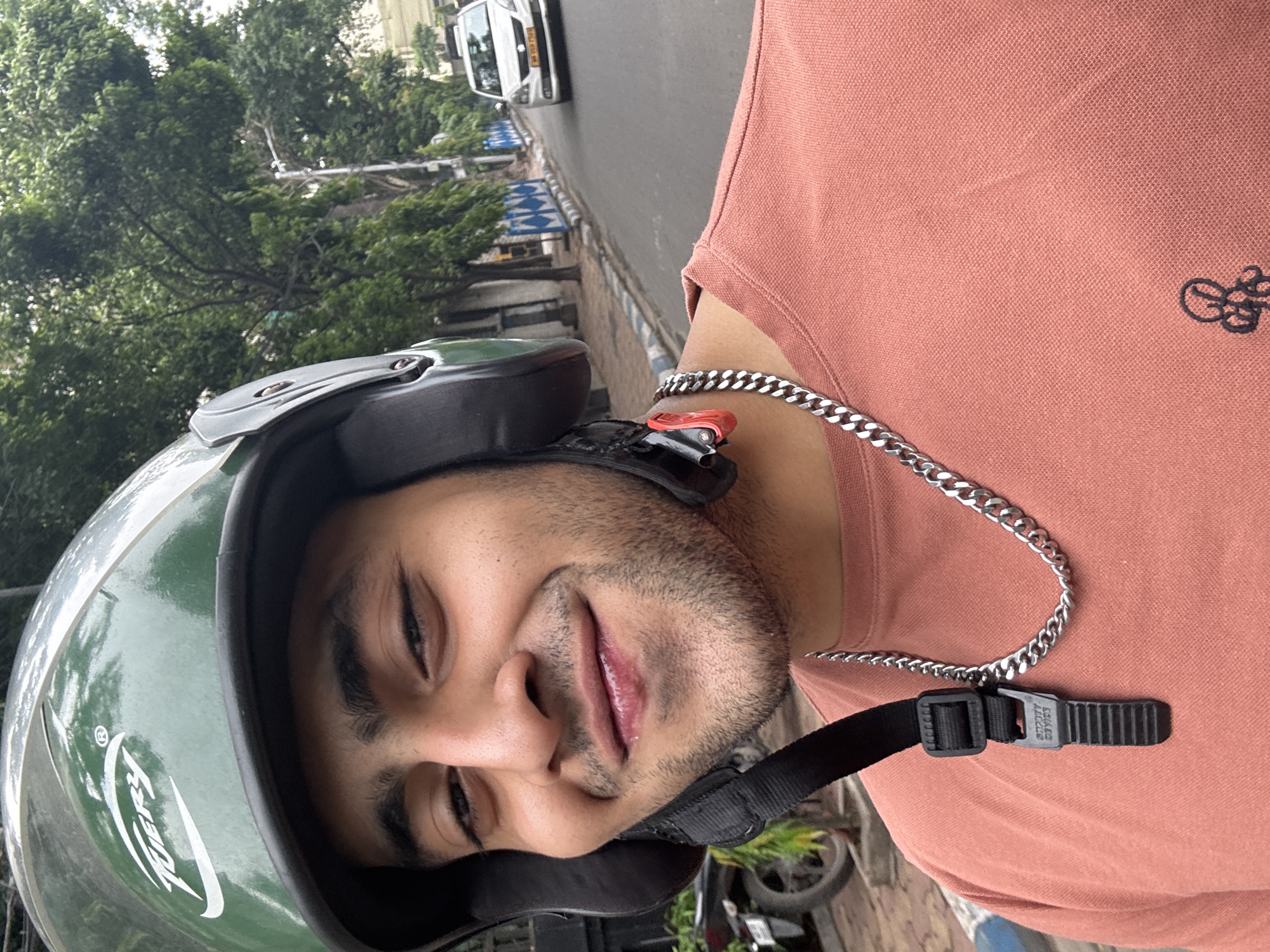 A man wearing a green helmet and a salmon-colored polo shirt is riding a vehicle on a street in Kolkata, India. He has a silver chain around his neck. The environment includes a street with a car and trees, with blue and white traffic barriers visible along the roadside. The image is taken at an angle, suggesting a selfie perspective while the man is moving.
