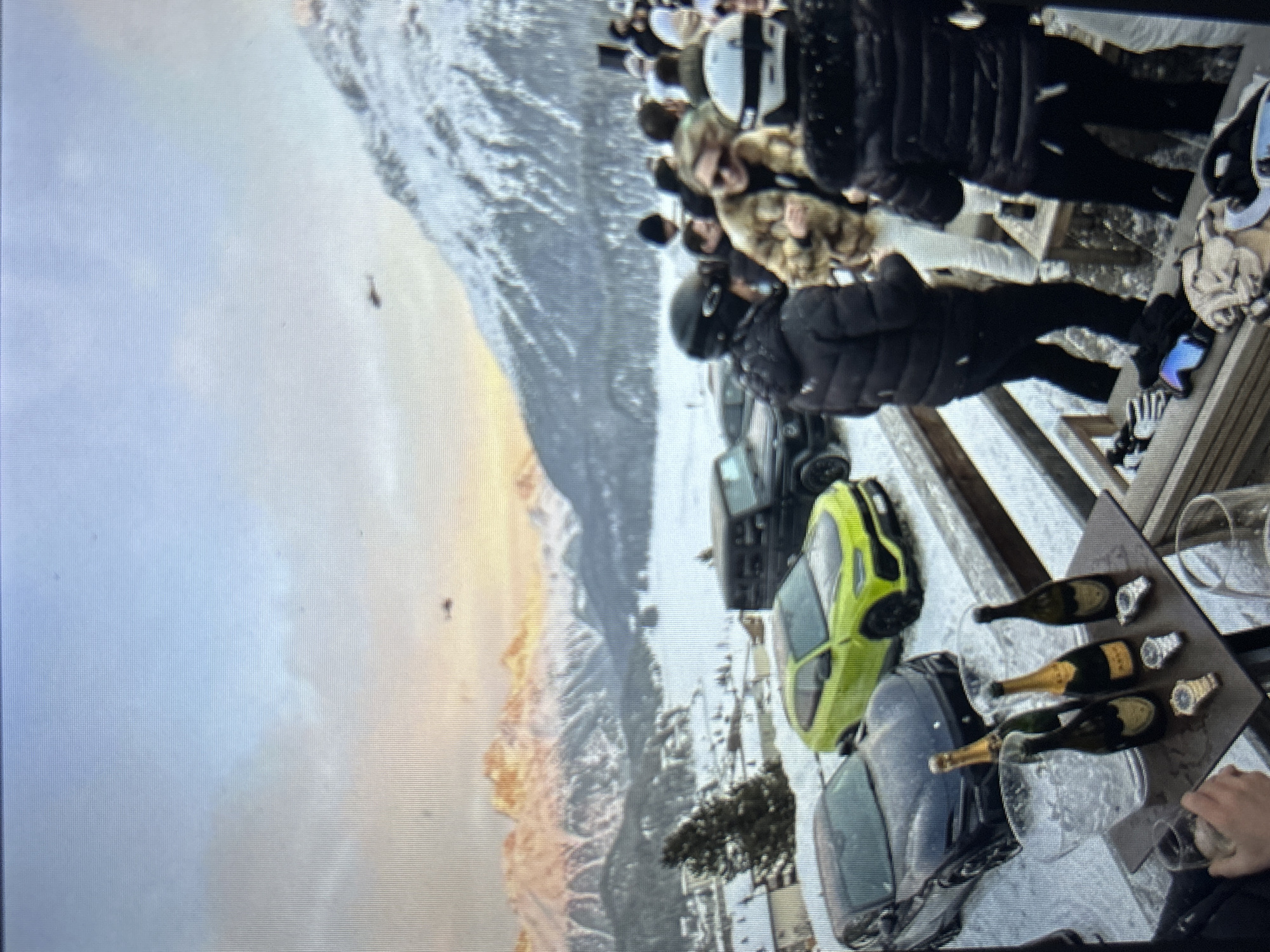 The image depicts a winter scene set in the city of Burgas, Bulgaria, at what appears to be a high-altitude outdoor terrace overlooking a vast, snow-covered mountainous landscape. The sky exhibits hues of blue, orange, and pink, suggesting either sunrise or sunset, illuminating distant snow-capped peaks. A large, frozen body of water is visible in the valley below, surrounded by snow-dusted terrain and scattered structures.
In the foreground, a wooden terrace is partially covered in snow. On a dark-colored table, a clear ice bucket holds ice, alongside five bottles of champagne; two of these bottles clearly display "Dom Pérignon" labels, while one has gold foil and two have dark green foil. Three luxury watches with metal bands – two silver-toned and one gold-toned – are meticulously arranged on the table next to the champagne. A person's hand is visible in the lower left, holding a clear glass containing a light-colored beverage. Ski goggles with blue lenses and black gloves are placed on a wooden railing.
Three luxury SUVs are parked on a snowy surface directly behind the terrace: a dark-colored Lamborghini Urus, a vibrant lime green Lamborghini Urus, and a dark-colored Mercedes-Benz G-Wagen further in the background.
A group of people are gathered on the terrace, most dressed in heavy winter clothing, including puffer jackets and ski helmets. One individual wears a light brown fur coat and sunglasses, smiling towards the right. Another person in a black puffer jacket and black ski helmet faces left, partially obscured. A third person, with their back to the viewer, wears a black puffer jacket with a fur-lined hood and a white ski helmet. The people appear to be socializing.
In the distant sky, at least three small, dark-colored helicopters are visible against the backdrop of the mountains and sky.