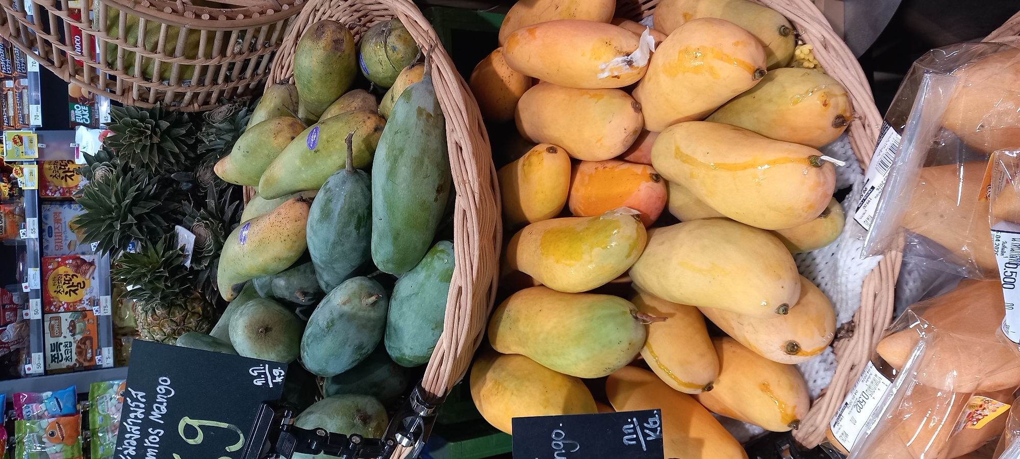 This image captures a vibrant section of a grocery store or market, likely in Moo 1, Thailand, featuring an abundant display of fresh mangoes and packaged goods.
In the foreground, two large, woven baskets are filled with mangoes. The basket on the left contains a mix of green and partially ripened yellow-green mangoes, with several spiky pineapples nestled behind them. A black pricing sign in front of this basket reads "ม่วงสามรัส" (Mang Sam Rus) and "mros Mango," indicating the variety, along with "ก.ก. / kg." The number '9' is partially visible on the sign. The basket on the right is overflowing with bright yellow and orange ripe mangoes, some exhibiting sticky residue or natural juices on their skin. Several of these ripe mangoes are individually packaged in clear plastic, with visible labels showing "น้ําหนักสุทธิ 0.502 กก." (Net weight 0.502 kg) and "วันที่ผลิต 6.04.2020" (Production date 06.04.2020) on one, and "0.500 กก" (0.500 kg) on another.
To the left of the fruit display, shelves are stocked with various packaged snacks and biscuits. Visible product names include "チョコチップ" (Choco Chip), "チョコ&コーヒービスケット" (Choco & Coffee Biscuit), "초코파이" (Choco Pie), "유자치즈케익" (Yuja Cheese Cake), and "EURO CAKE." Prices in Thai Baht are displayed on shelf labels, such as "70," "55," "50," "175," and "45," with some "Sale" signs. The scene portrays a lively retail environment, offering both fresh local produce and a variety of international convenience items.