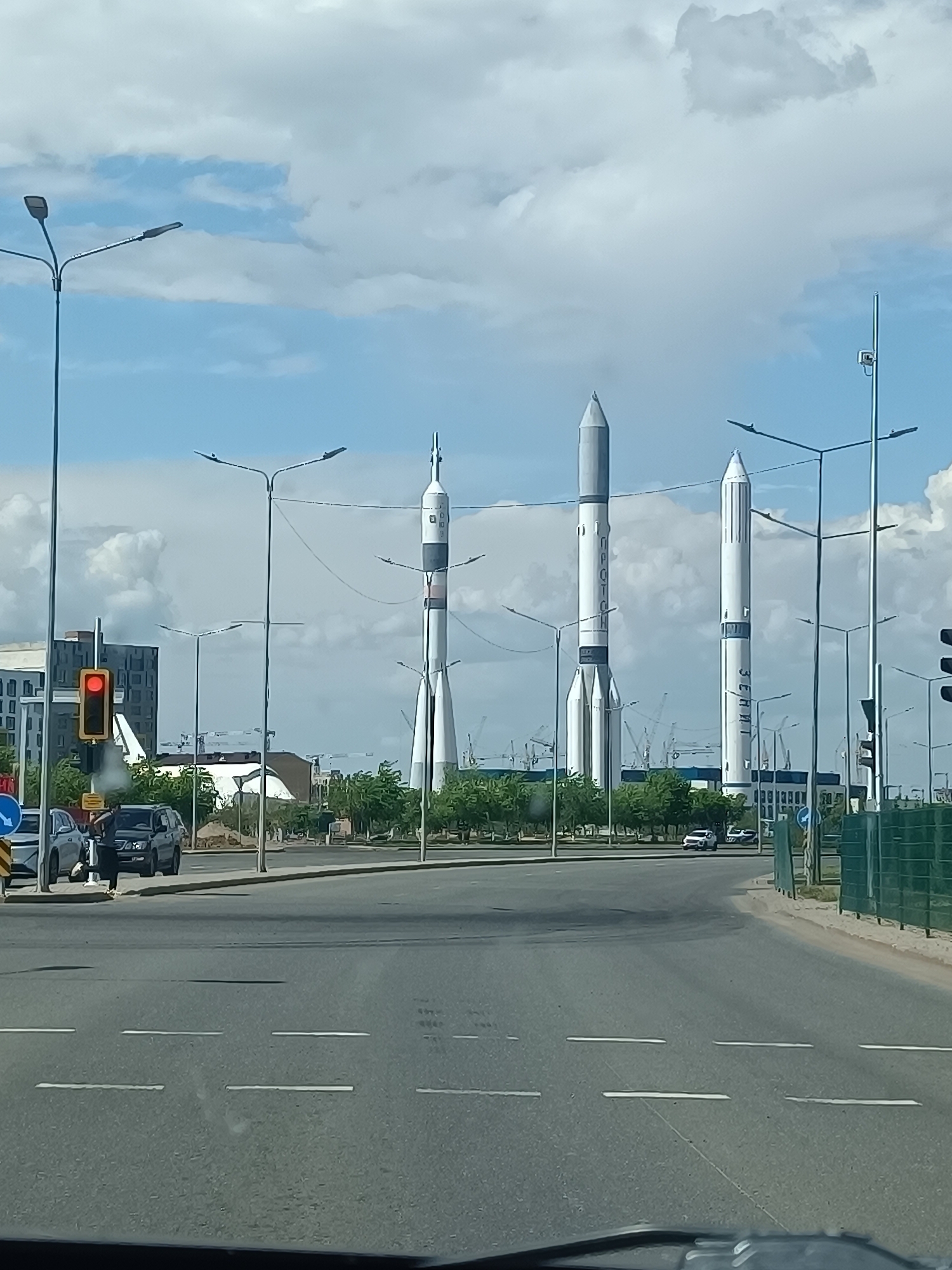 The image shows a street view in Astana, Kazakhstan, featuring three large rocket monuments. The rockets, from left to right, appear to be replicas of the Soyuz, Proton, and Zenit rockets. The scene is viewed from within a vehicle, showing a wide street with multiple lanes and white dividing lines. On the left side of the road, a traffic light is visible, showing a red light. Cars are parked on the roadside and a person is standing next to one of the vehicles. The rockets are situated in a park-like area, with numerous trees and lamp posts between the street and the monuments. Construction cranes are visible in the background. The sky is partly cloudy.