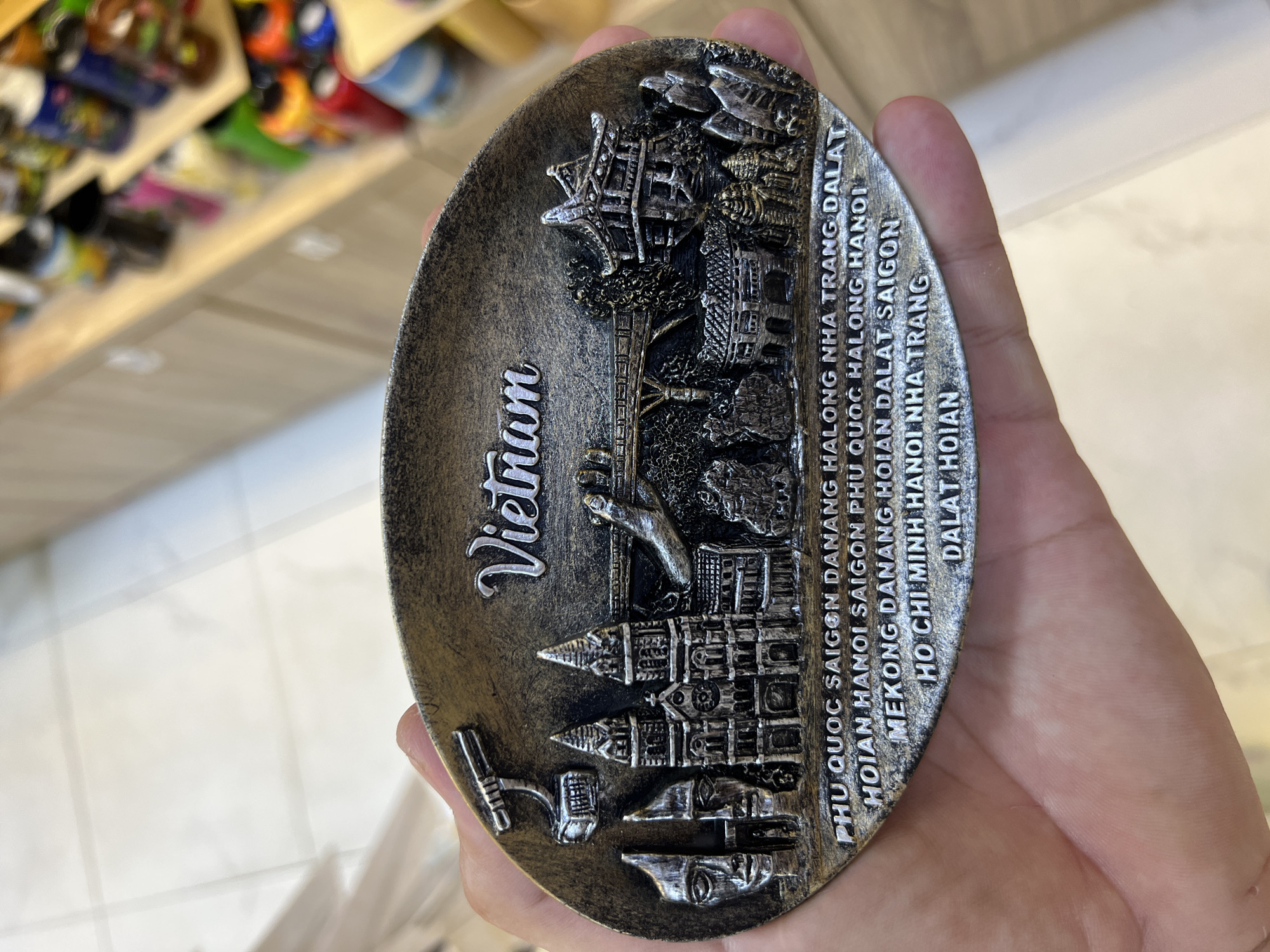 A hand holds a metallic souvenir plate featuring a relief of Vietnamese landmarks and city names. The plate is oval-shaped and has a bronze and silver finish. The word "Vietnam" is prominently displayed in silver lettering at the top. Below, a detailed cityscape includes architectural styles reminiscent of Hanoi and Ho Chi Minh City, a bridge, and a mountainous landscape. The lower portion of the plate is inscribed with a list of Vietnamese cities: "PHU QUOC SAIGON DANANG HALONG NHA TRANG DALAT HOIAN HANOI SAIGON PHU QUOC HALONG HANOI MEKONG DANANG HOIAN DALAT SAIGON HO CHI MINH HANOI NHA TRANG DALAT HOIAN". The background shows blurred shelves with various products, suggesting the souvenir is in a shop. The hand holding the plate is light-skinned. There is no indication of the specific location within Nha Trang, Vietnam.