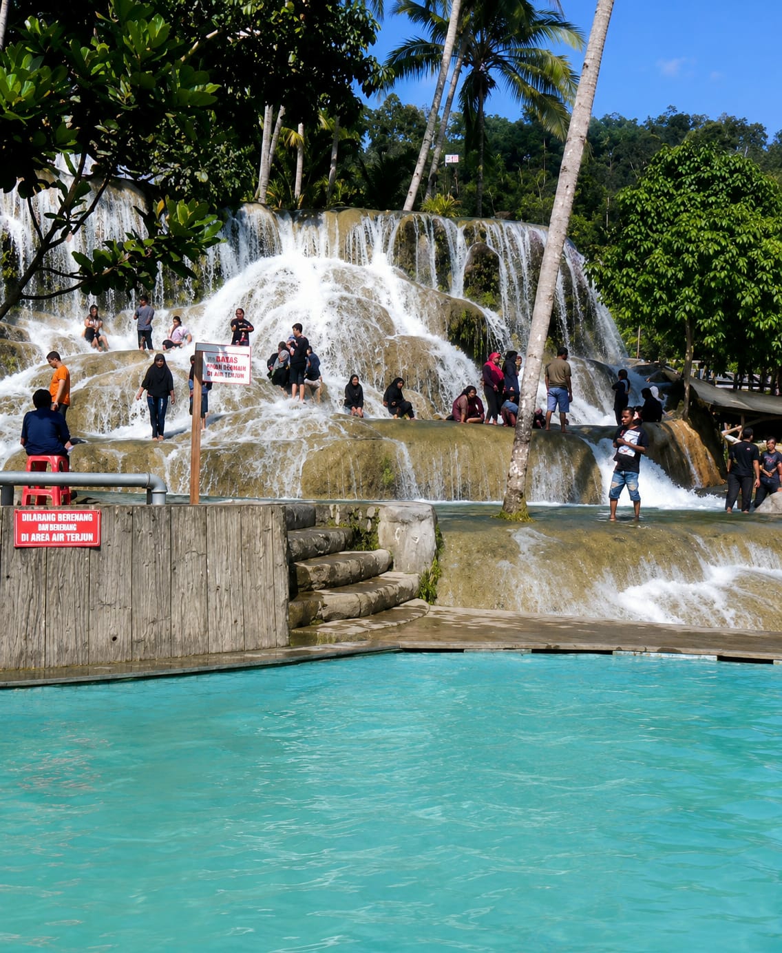 The image shows a tiered waterfall with people interacting with the water. In the foreground, a bright turquoise swimming pool is visible, with a concrete edge and a wooden barrier. A red sign is attached to the wooden barrier, with text in Indonesian that reads "DILARANG BERENANG DAN BERMAIN DI AREA AIR TERJUN" which translates to "SWIMMING AND PLAYING ARE PROHIBITED IN THE WATERFALL AREA".
Behind the pool, the waterfall cascades down a rocky, multi-layered formation. Several individuals are seated on the rocks, some near the water's edge, while others are standing or walking on the wet surfaces. A prominent palm tree trunk is positioned in front of the waterfall. Trees and foliage surround the top of the waterfall, with a clear blue sky visible above. The scene appears to be a public recreational area, likely within the city of Silaiyajae, Indonesia, based on the provided context, featuring a natural waterfall integrated with man-made swimming facilities.