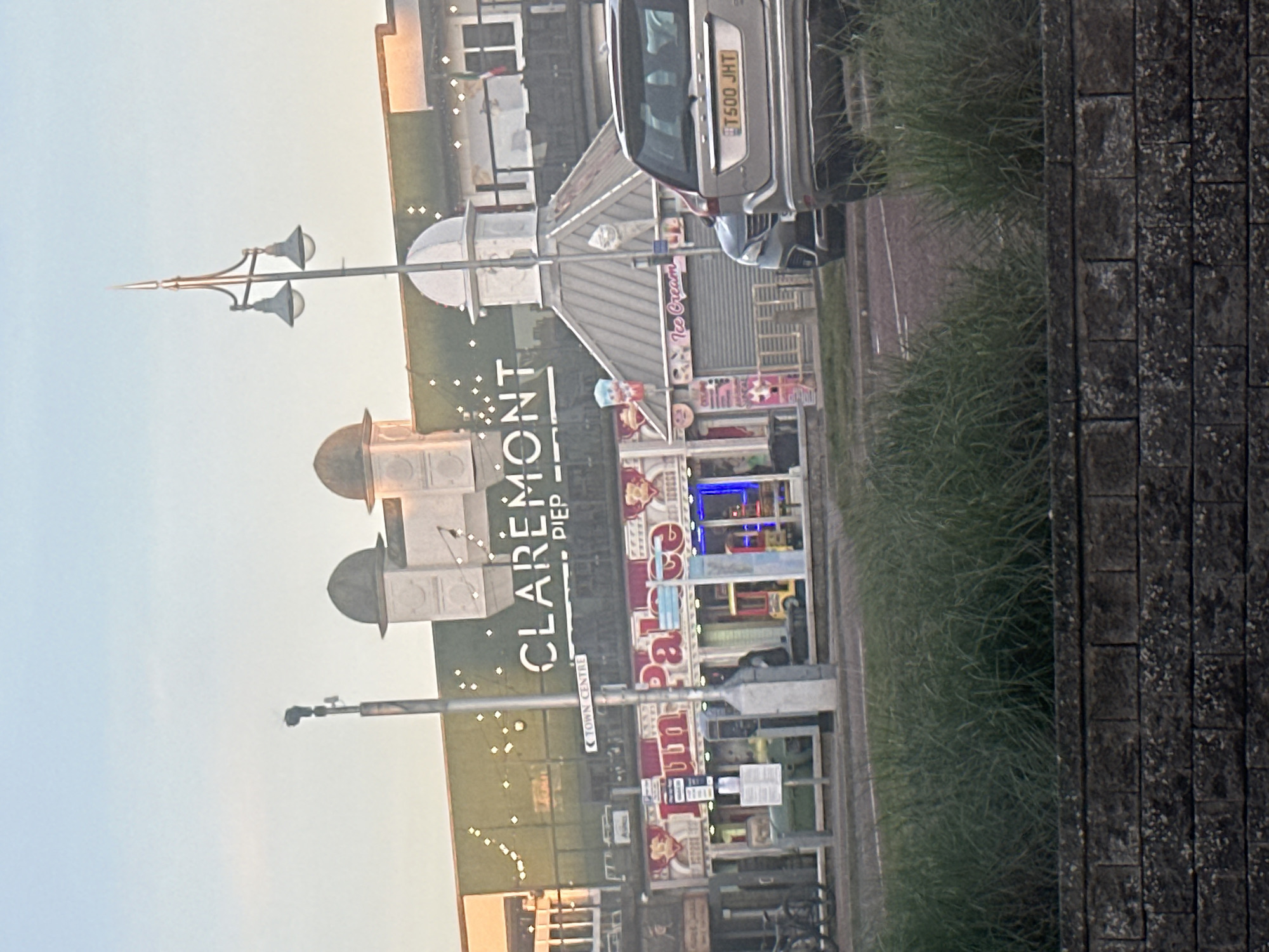 The image depicts a view of Claremont Pier in Lowestoft, East Suffolk, United Kingdom. The pier's architecture, featuring decorative domes, is visible, alongside signage that reads "CLAREMONT PIER" and "Ice Cream." The pier's amusements and retail fronts can be seen. A street lamp stands in the foreground, and a parked car with license plate "T500 JHT" is also visible near the pier's entrance. Grass and a brick wall border the scene on the right. The sky is light-colored, suggesting daytime.