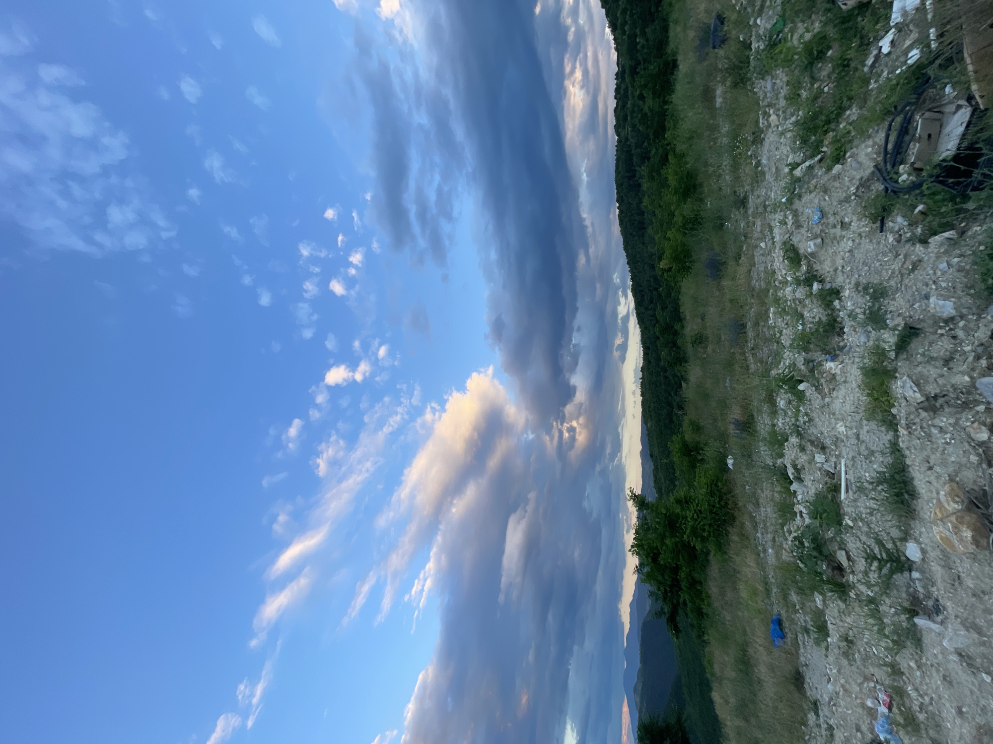 The image captures a landscape view from a location identified as Bogolin, Bulgaria. The foreground is a rugged, rocky terrain with scattered debris, including plastic bags, wrappers, and discarded items. Beyond this disturbed ground, a grassy field transitions into a densely wooded hillside. In the distance, a series of rolling, forested hills are visible against a sky filled with a mix of clear blue and dramatic clouds. The clouds are wispy and elongated in some areas, tinged with the warm hues of sunset or sunrise, while others appear darker and more consolidated. The overall impression is of a natural environment marred by human-generated waste, with the broader landscape suggesting a rural or semi-rural setting on the outskirts of the town.