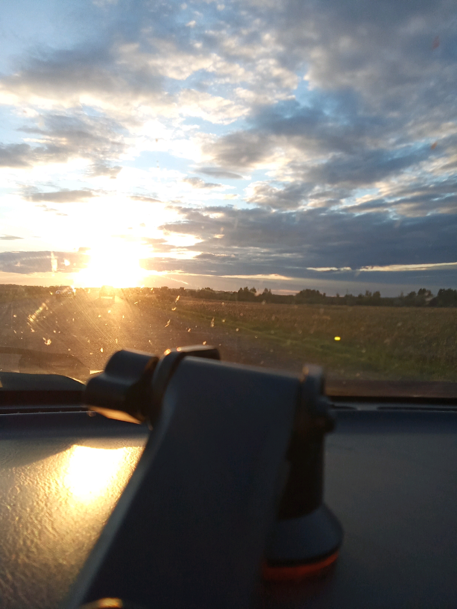 The image appears to be taken from inside a vehicle, looking forward through the windshield. The foreground is dominated by the dashboard and a dark-colored object, possibly a phone mount, partially obscuring the view. The windshield is smudged with dirt or dust, creating a hazy effect, particularly around the sun.
The main focus of the image is the sky, which is filled with a dramatic sunset. The sun is a bright, intense light source, low on the horizon, casting a warm, golden glow across the scene. Streaks of light refract through the dirty windshield. The sky itself is a mix of blue and grey clouds, with the sun's rays breaking through in several places, creating a vibrant and dynamic atmosphere.
Beyond the windshield, a flat, rural landscape stretches out to the horizon. It appears to be agricultural land, possibly fields of crops or harvested land, with some trees or vegetation visible in the distance. The road leading towards the horizon is illuminated by the setting sun, and there are faint outlines of vehicles on the road ahead.
The overall impression is one of a journey at sunset, with the natural beauty of the sky juxtaposed against the mundane interior of a vehicle. The image captures the atmospheric conditions and the visual experience of traveling during golden hour. The location is identified as Рощин ауылдық округі, Kazakhstan.