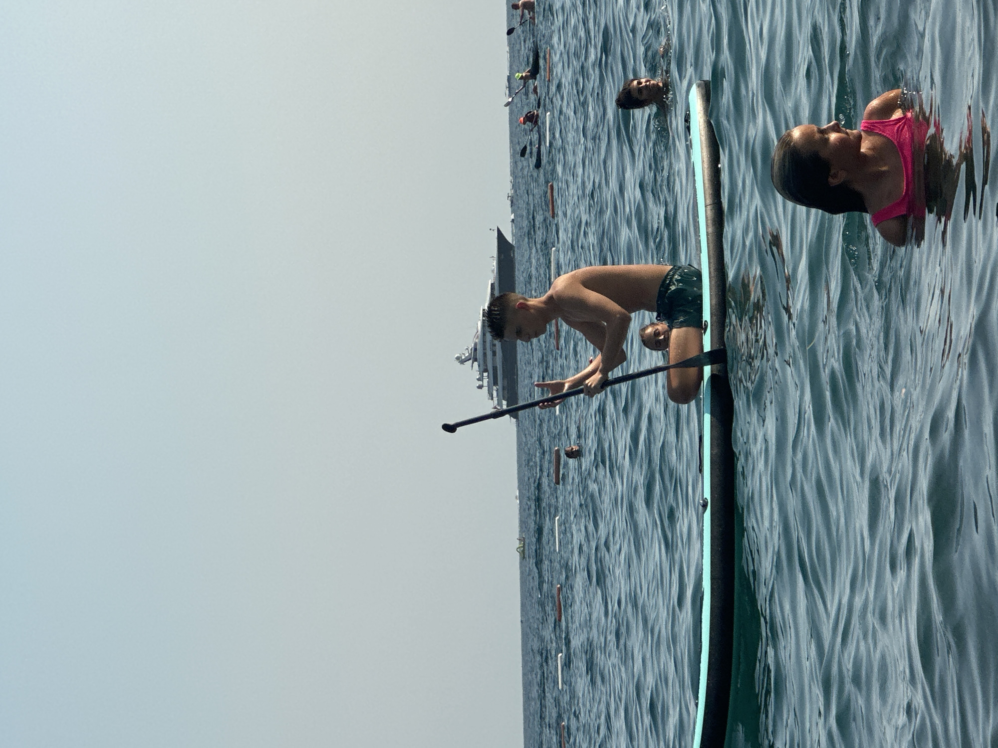 A wide shot captures a group of people enjoying a sunny day on the water in Boreti, Montenegro. In the foreground, a young man is seated on a paddleboard, holding a paddle. To his right, a young girl in a pink swimsuit is swimming in the sea. Behind them, several other individuals are visible in the water and on other paddleboards. In the background, a large yacht sails on the horizon, under a clear, pale blue sky. The water is a deep blue, with gentle ripples. Bouy lines are visible in the middle ground, marking a swimming area.