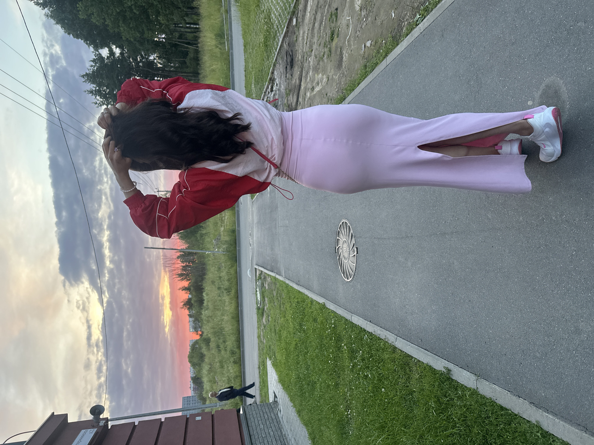 A rear view of a woman with long, dark hair stands on a paved walkway in Murino, Russia. She is wearing a light pink maxi skirt with a high slit and a red and white jacket. Her hands are raised, adjusting her hair. She has a small pink purse slung across her body. To her left, a man in a dark suit walks away from the camera on a parallel paved path. The walkway is bordered by a grassy area and then a road. In the background, a vibrant pink and orange sunset illuminates the sky above a line of trees and buildings. A brown brick building with a speaker attached to a pole is visible to the left of the frame. Power lines crisscross the sky. A manhole cover is visible on the walkway.
