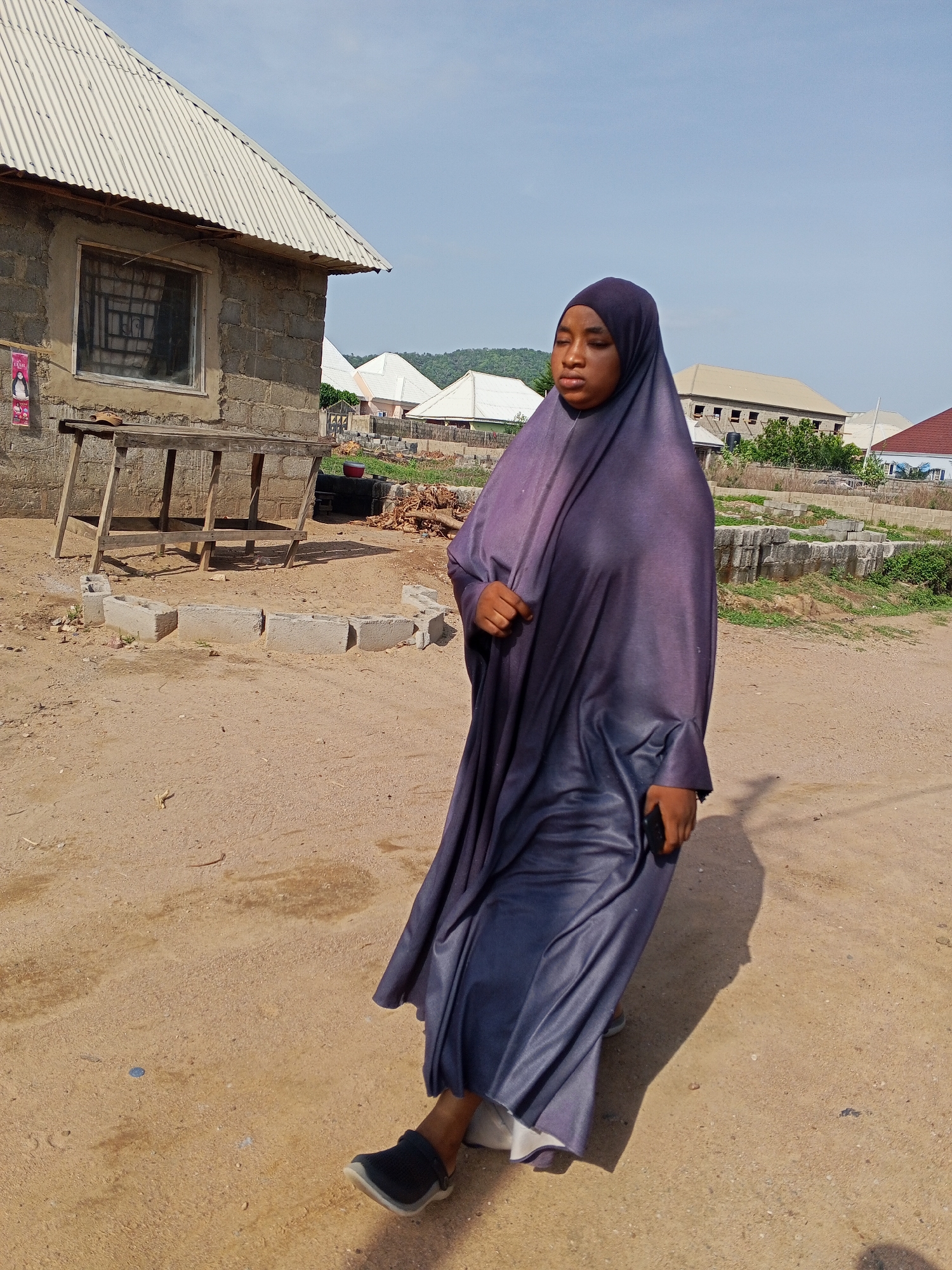 A full-body shot captures a young individual, likely female, walking outdoors in Adonkolo, Nigeria. She is dressed in a flowing, dark purple or blue abaya/jilbab and a matching hijab that covers her head and shoulders. She looks towards her left (viewer's right) with a neutral expression. In her right hand, she holds a black smartphone, and she wears dark, slip-on shoes on her bare feet.
The setting is a dusty, unpaved area, characteristic of a rural or developing urban environment. To the left, a simple, unfinished building constructed from concrete blocks stands prominently, featuring a corrugated metal roof and a single window secured with grilles. A rustic wooden table or stand is positioned next to this building. On the ground in front of it, concrete blocks are arranged in a rough semi-circle. Further in the background, other buildings with similar roofing styles are visible, interspersed with green foliage and distant hills under a clear, bright blue sky. The sunlight is strong, casting distinct shadows, indicating daytime. A faded poster with a woman's image and indistinct text is affixed to the wall of the concrete block building. The ground is covered in dry earth, with scattered debris and construction materials visible in the mid-ground and background.