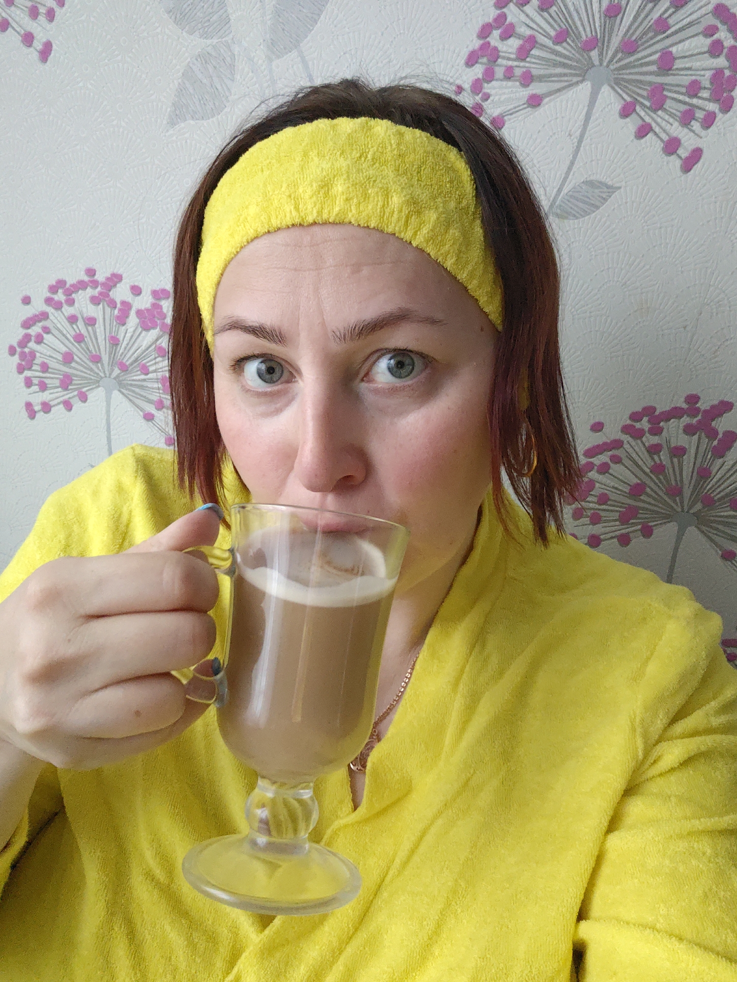 A woman with brown hair and blue eyes is drinking from a clear glass filled with a tan-colored beverage with a foamy top. She is wearing a yellow terrycloth headband and a matching yellow robe or jacket. She has a gold necklace and small gold hoop earrings. Her nails are painted a light blue. The background is a wall with a patterned wallpaper featuring abstract floral designs in shades of pink and gray. The location is unknown, and there is no evidence to support that it is in the city of Markovskii, Russia.