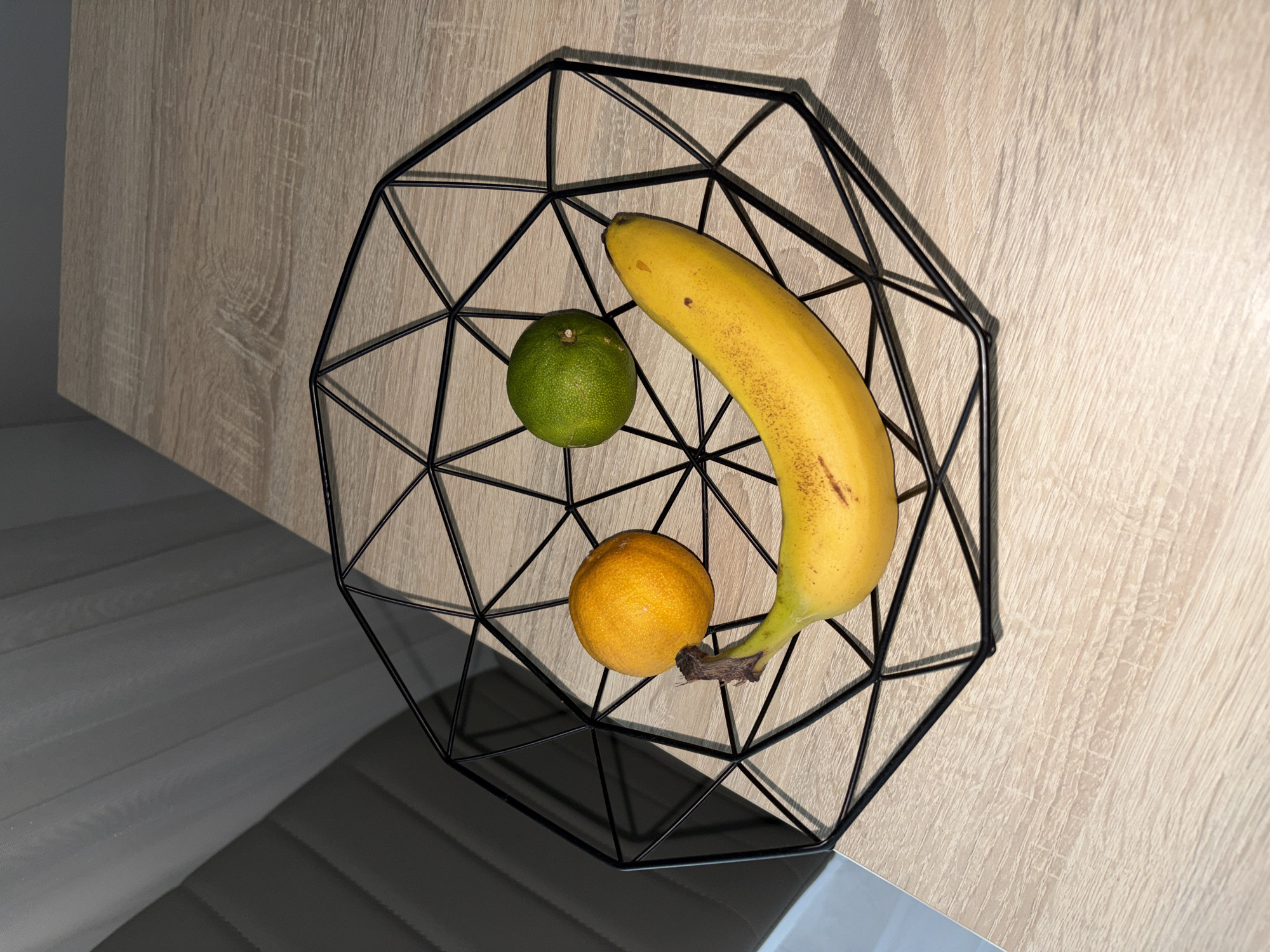 A geometric black wire fruit basket sits on a light-colored wooden surface. Inside the basket are a yellow banana, an orange, and a lime. The fruits are arranged to resemble a smiley face, with the banana forming the mouth, the orange and lime as eyes. The background consists of a gray, textured surface, possibly blinds or a wall covering. The scene appears to be an indoor still life. No specific location in Nitra, Slovakia can be determined from the provided image.