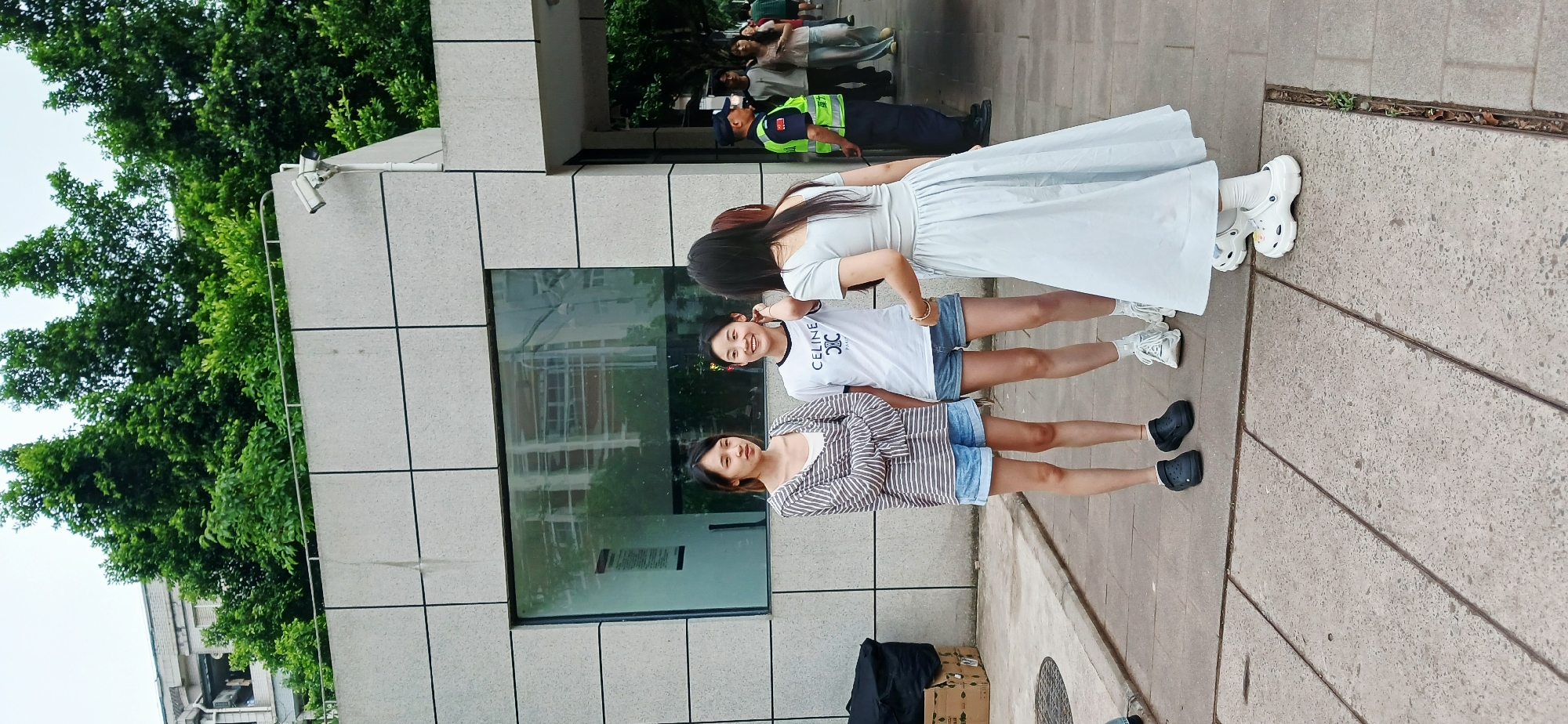 Three women are standing on a sidewalk in the city of Chongqing High-tech Zone, China, next to a modern building with square, light-colored facade panels. The building has a large glass window reflecting the environment. To the left of the building, there is lush greenery. One woman is wearing a long, light-colored dress and white sneakers. Another woman is wearing a white t-shirt with the word "CELINE" on it and denim shorts with white sneakers. The third woman is wearing a striped top, denim shorts, and black shoes. To the right of the women is concrete pavement. In the background are more people, including a person wearing a security uniform with a yellow vest. There are also some packages on the ground near the bottom of the frame. There is a security camera on the building.