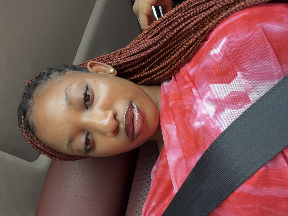 The scene depicts a female individual seated within a vehicle in Ikota, Nigeria. Her upper torso and head are visible in a close-up, angled shot. She has medium brown skin, and her hair is styled in numerous small, reddish-brown box braids that extend over her shoulders. Her eyes are partially closed or downcast. Her lips are coated with a dark red, glossy substance. A small, spherical gold stud earring is visible on her right earlobe. She is wearing a top or dress made of a sheer, fine-mesh fabric, exhibiting a red, pink, and white tie-dye or mottled pattern. A dark grey seatbelt is fastened across her chest, running diagonally from the upper left to the lower right of the frame. The immediate background shows a dark reddish-brown upholstered surface to her left and a lighter, possibly grey or beige, ceiling or upper interior panel to her right. A metallic-colored object is faintly visible on the far right.