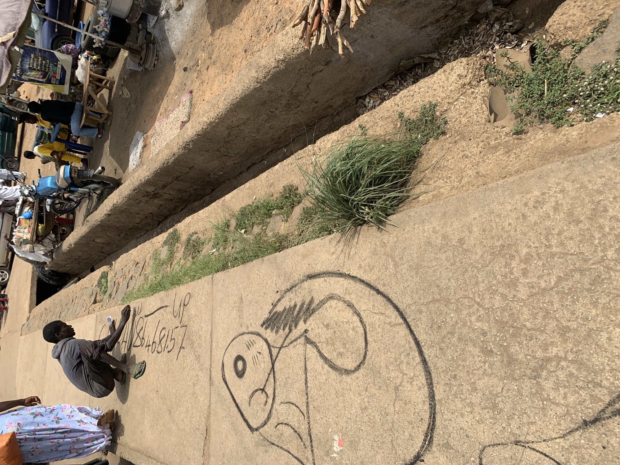 The image depicts a man kneeling on a concrete sidewalk in what appears to be a street in Rafin Par, Nigeria. He is writing numbers in chalk next to a crude drawing of a head. The numbers are written in black chalk and are approximately 86468157. The sidewalk abuts a raised concrete drainage ditch. The ditch is partially overgrown with grass and weeds. In the background on the other side of the ditch, there is evidence of commercial activity with people seated on chairs and motorcycles parked nearby.