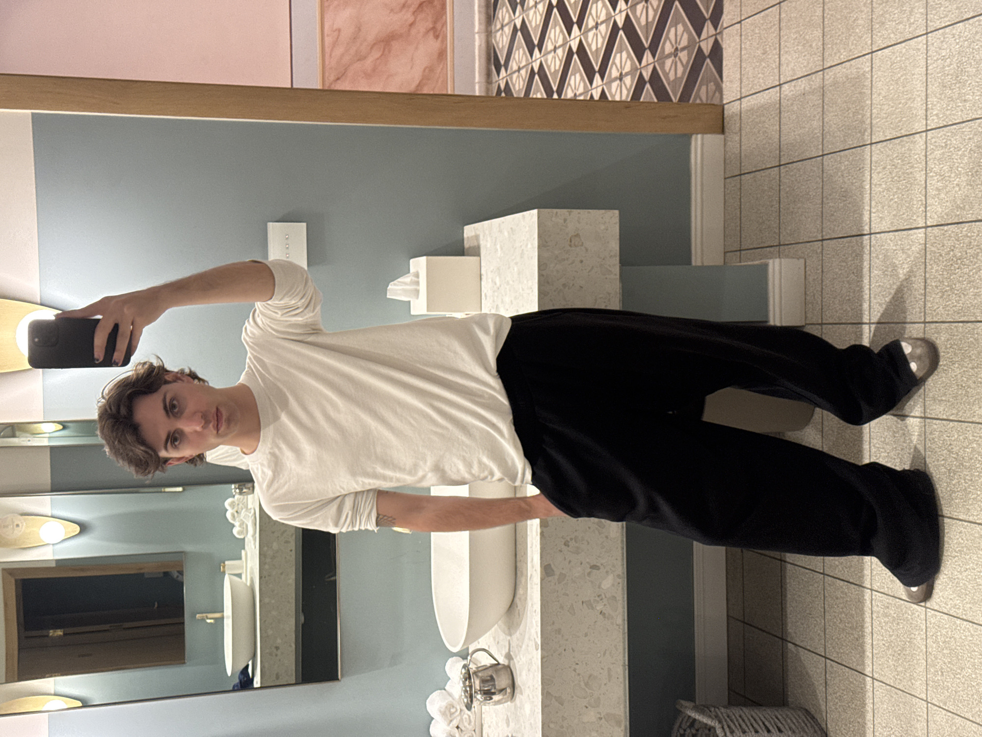 A young man stands in a bathroom, posing for a selfie in the mirror. He is wearing a white long-sleeved shirt and black pants with silver-colored shoes. The bathroom features a modern design, including light blue walls, a countertop with a white sink, and tile flooring. The background shows geometric patterns on the wall, possibly indicating a stylish or boutique hotel location. The location is likely not in Tel-Aviv, Israel, as there are no specific visual cues pointing to that location.