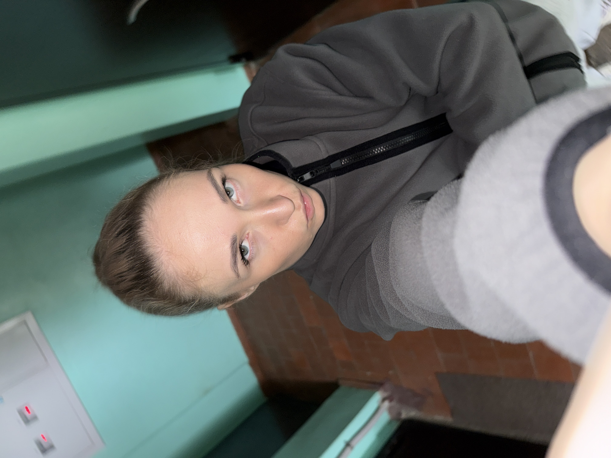 The image is a close-up, tilted portrait of a young woman, likely a selfie, taken indoors. The woman, the primary subject, is positioned centrally in the frame. She has light skin, light-colored eyes (appearing green or blue), and light brown hair pulled back, revealing her forehead. Her expression is neutral, with her lips slightly parted. She is wearing a dark grey, possibly fleece, jacket with a prominent black zipper running down the front. Part of a lighter grey sleeve is visible on the right side of the image, suggesting a layered outfit or a two-toned garment.
The background reveals an indoor setting, possibly a hallway or stairwell. On the left side, there is a flat wall painted in a light turquoise or teal color. On the right, a reddish-brown brick wall or surface is visible. In the bottom-left corner, a white rectangular panel with two small red indicator lights, resembling an electrical box or intercom, is partially visible. The floor area below the brick wall appears dark. The entire image is strongly tilted to the right, creating a dynamic, almost sideways perspective. The lighting is diffused and even, suggesting an indoor environment without strong natural light. No specific activity other than posing for the photograph is depicted. There is no visible text in the image. The architectural elements are consistent with typical interior building designs found in residential areas, aligning with the provided location context of Chaikovsky, Russia.
