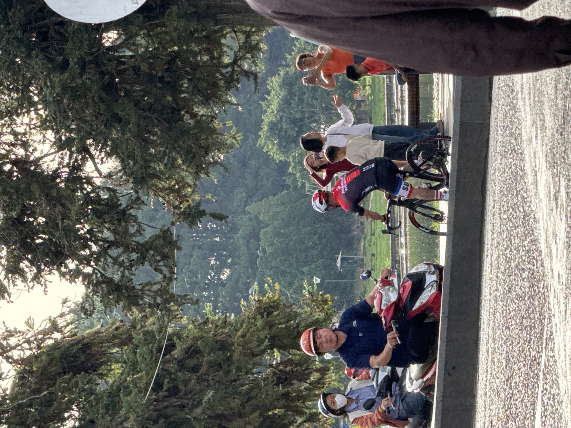 The image depicts a scene in Da Lat, Vietnam, likely near Xuan Huong Lake, characterized by lush greenery and a bright, overcast sky. In the foreground, a person rides a red scooter with another person behind them wearing a face mask and a white top. To the right, a cyclist in racing attire is paused, with several onlookers observing. A woman in an orange top is taking a photograph with her phone. Behind them, more individuals are gathered, some appearing to be tourists. The environment suggests a public park or recreational area, with trees and bodies of water visible in the background, indicative of the picturesque landscapes of Da Lat.