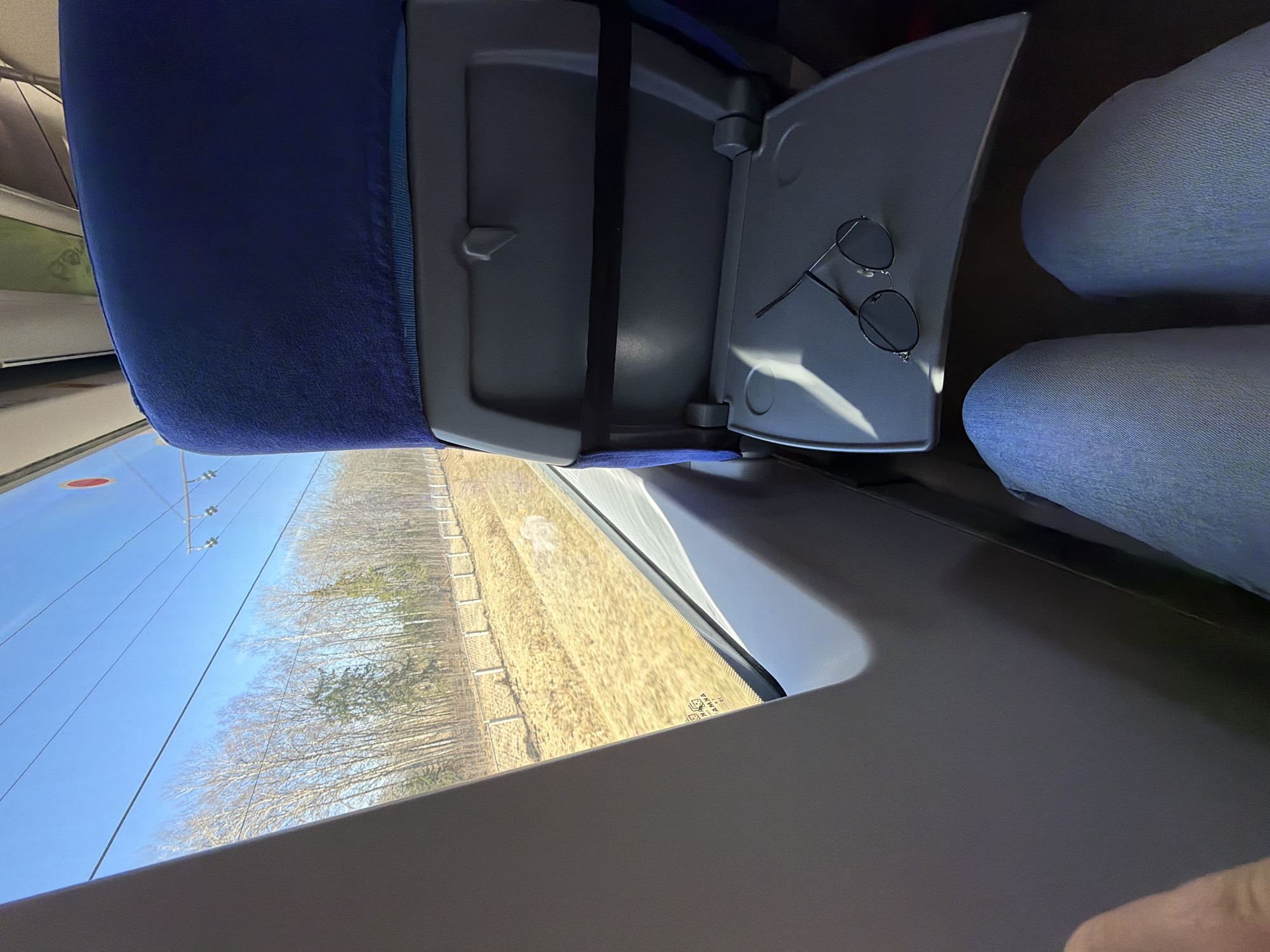 The image depicts an interior view from a train, likely in motion. The camera's perspective is from an overhead angle, focusing on a blue upholstered train seat, a folded-down gray tray table, and a window displaying a landscape. A pair of round sunglasses rests on the tray table. The viewer can see two people sitting in the seats to the right. The window reveals a clear blue sky, trees with bare branches, and a field with yellow grass. The location is Splav Massiva Babino, Russia.