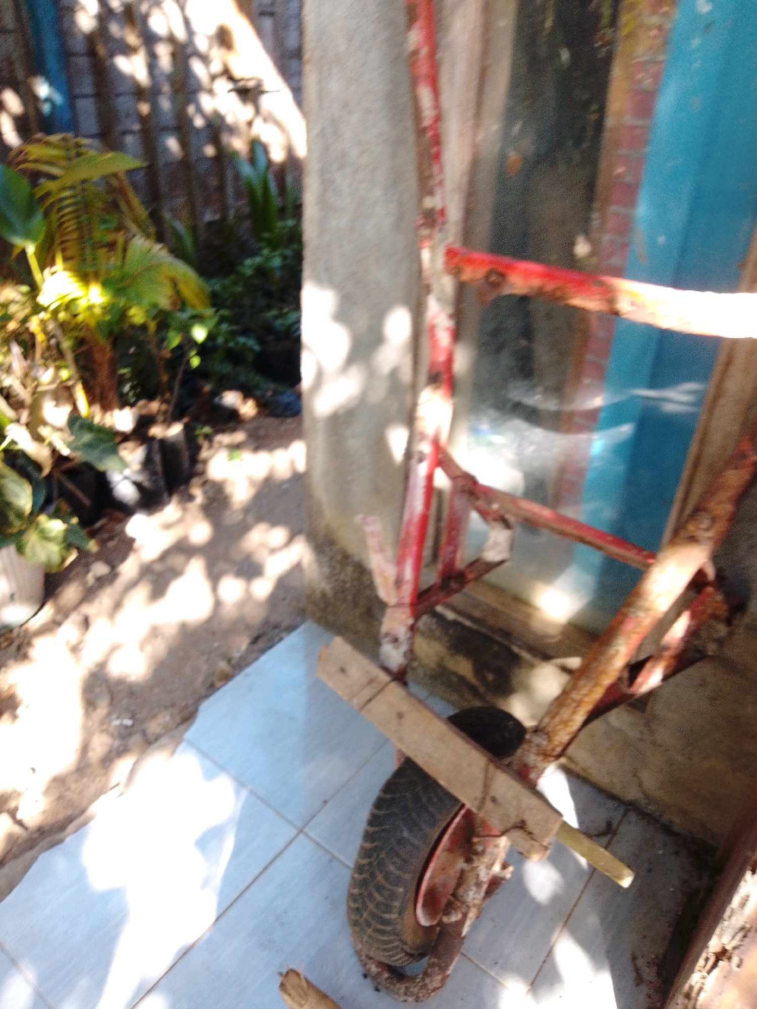 In Cikal, Indonesia, an outdoor scene is depicted with a wheelbarrow as the primary subject. The wheelbarrow, made of weathered red metal, is positioned on a tiled surface. It has a single black tire with a red rim. A wooden plank is attached to the frame of the wheelbarrow, extending outwards. To the left of the wheelbarrow, lush green plants are visible, some growing in pots. In the background, a brick wall with shadows cast upon it suggests an overcast or dappled sunlight condition. A concrete pillar stands to the right of the wheelbarrow. The overall environment appears to be a yard or outdoor storage area, possibly adjacent to a building.