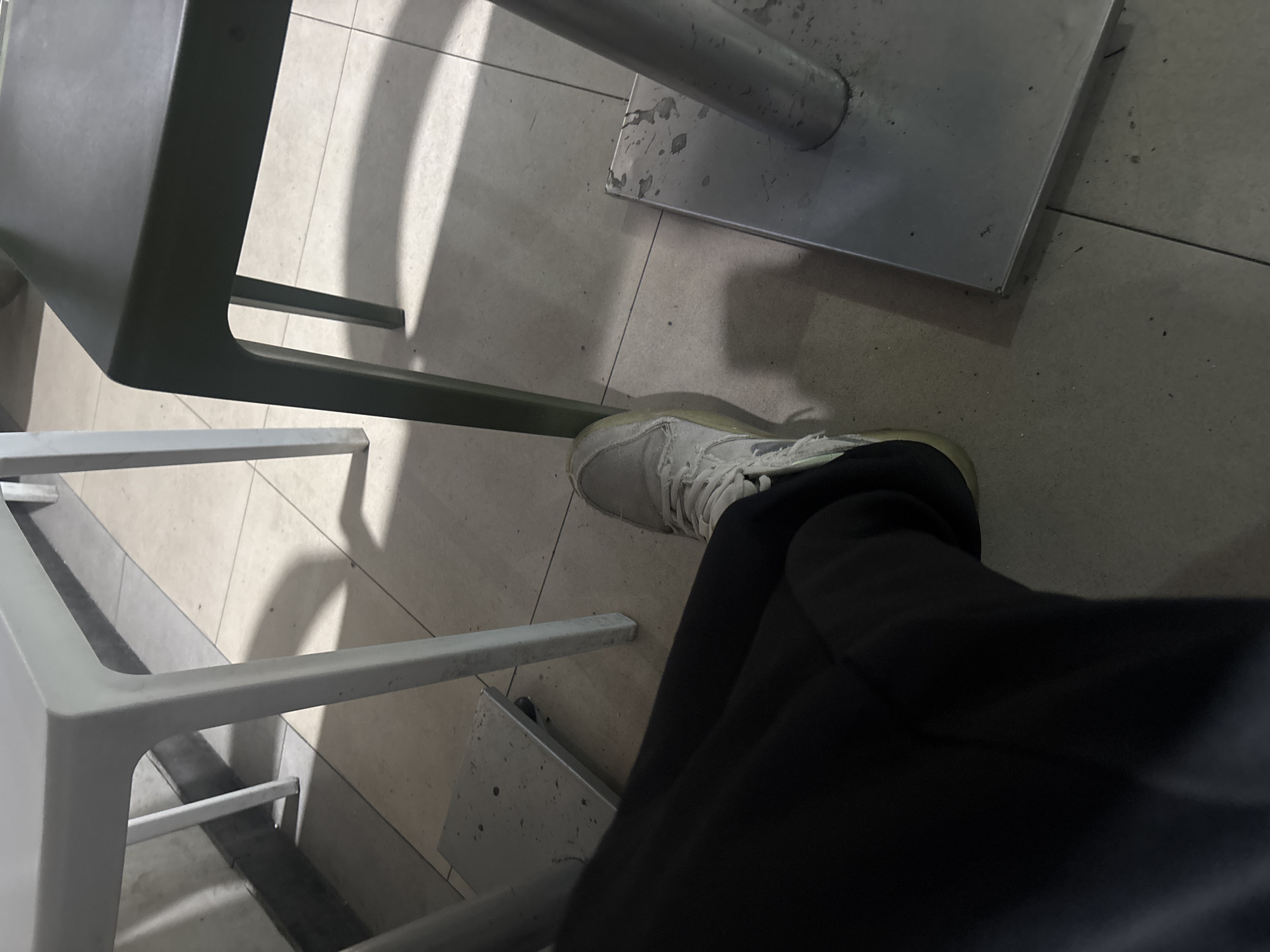 The image provides a low-angle, close-up view of a person's leg and foot within an indoor setting, likely a public area such as a cafe or waiting room in Yerevan, Armenia.
The scene focuses on the tiled floor and various metal furniture components. The floor consists of light-colored, possibly off-white or light grey, square tiles with darker grout lines, showing minor dirt and scuffs. A person's left leg, clad in black, relaxed-fit pants, is visible. Their foot wears a light grey or off-white sneaker with white laces and a distinct yellowish or light green sole. The person appears to be seated, with their leg extended slightly forward.
To the left, a sturdy, dark grey or greenish metal chair or stool leg forms a rounded U-shape, casting a strong, elongated shadow on the floor. Further down and behind it, thinner, lighter-colored rectangular metal bars are visible, which could be part of another chair or table's support structure. To the right, a square or rectangular metal base plate, likely for a table or stand, rests on the floor. This base plate is metallic grey and exhibits numerous small dark splatters and general wear.
The lighting appears to be artificial, creating well-defined shadows from the furniture on the tiled surface. There is no visible text in the image, and no specific activity other than the person sitting is evident.