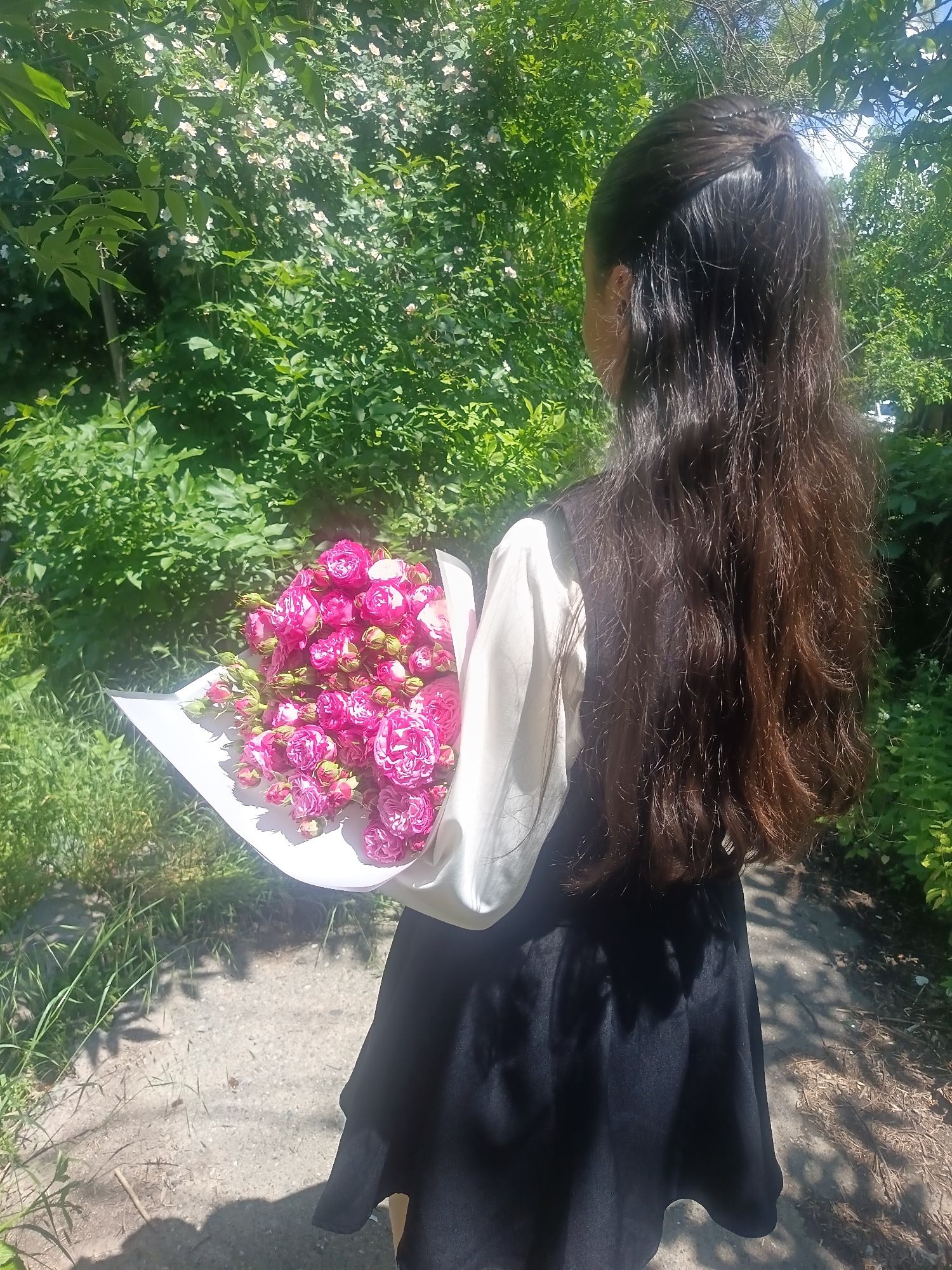 A young person stands facing away from the camera, holding a bouquet of pink roses. They have long, dark brown hair pulled back and are wearing a white blouse and a black skirt. The background is lush and green, with trees and foliage, suggesting an outdoor setting. Sunlight filters through the leaves, creating dappled shadows on the ground. The location is outdoors, in a naturalistic environment, with a path or walkway visible in the foreground.