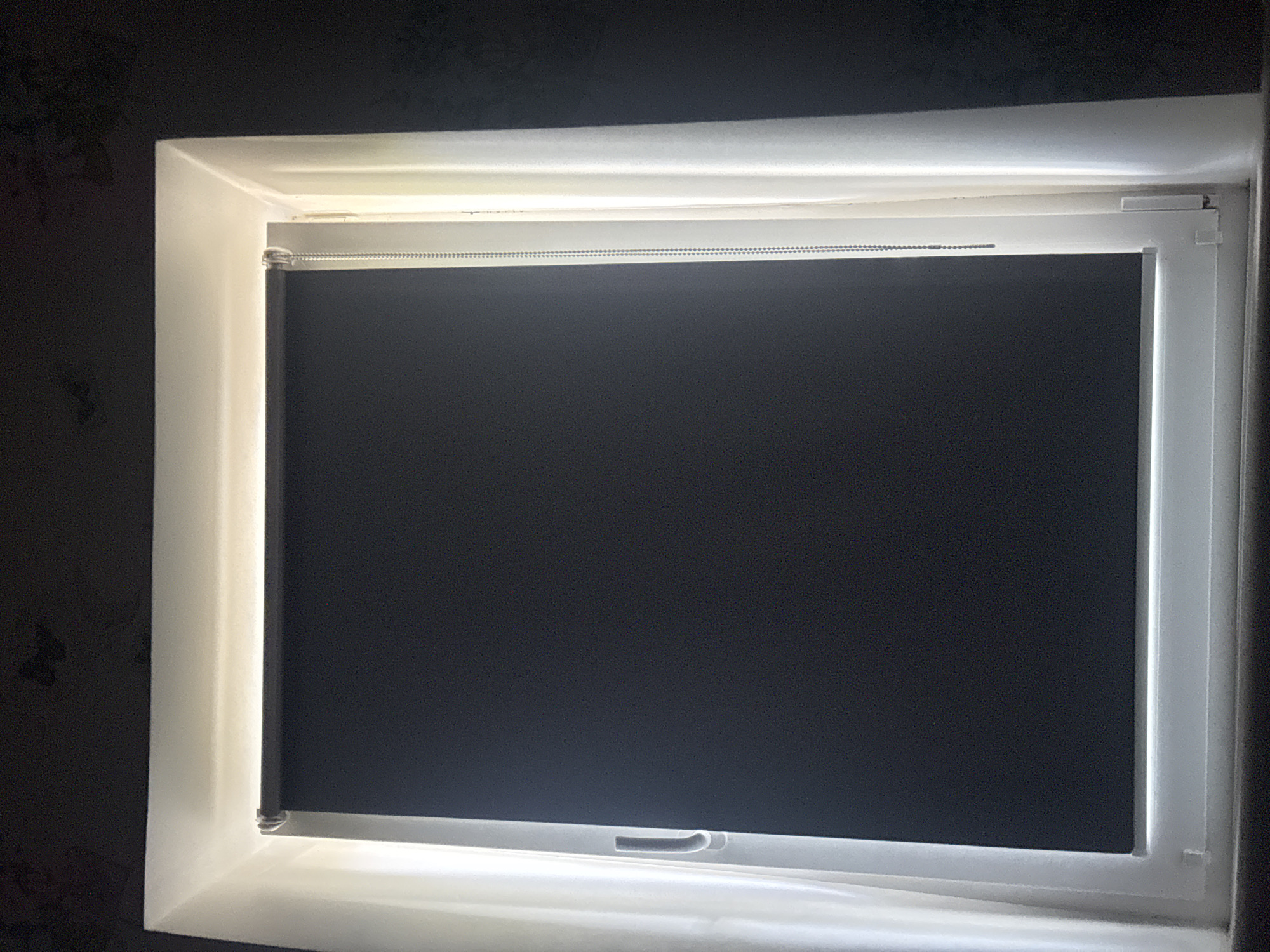 The image shows a closed, dark roller blind inside a white window frame. The window is illuminated from the outside by a bright light, creating a halo effect around the edges of the frame. A beaded pull cord for the blind is visible on the right side. The background is dark and appears to be a wall with a subtle floral pattern. The scene is indoors, likely a room in a residential or commercial building. No people are visible. The location in Kremenchuk, Ukraine, is not discernable from the image itself.