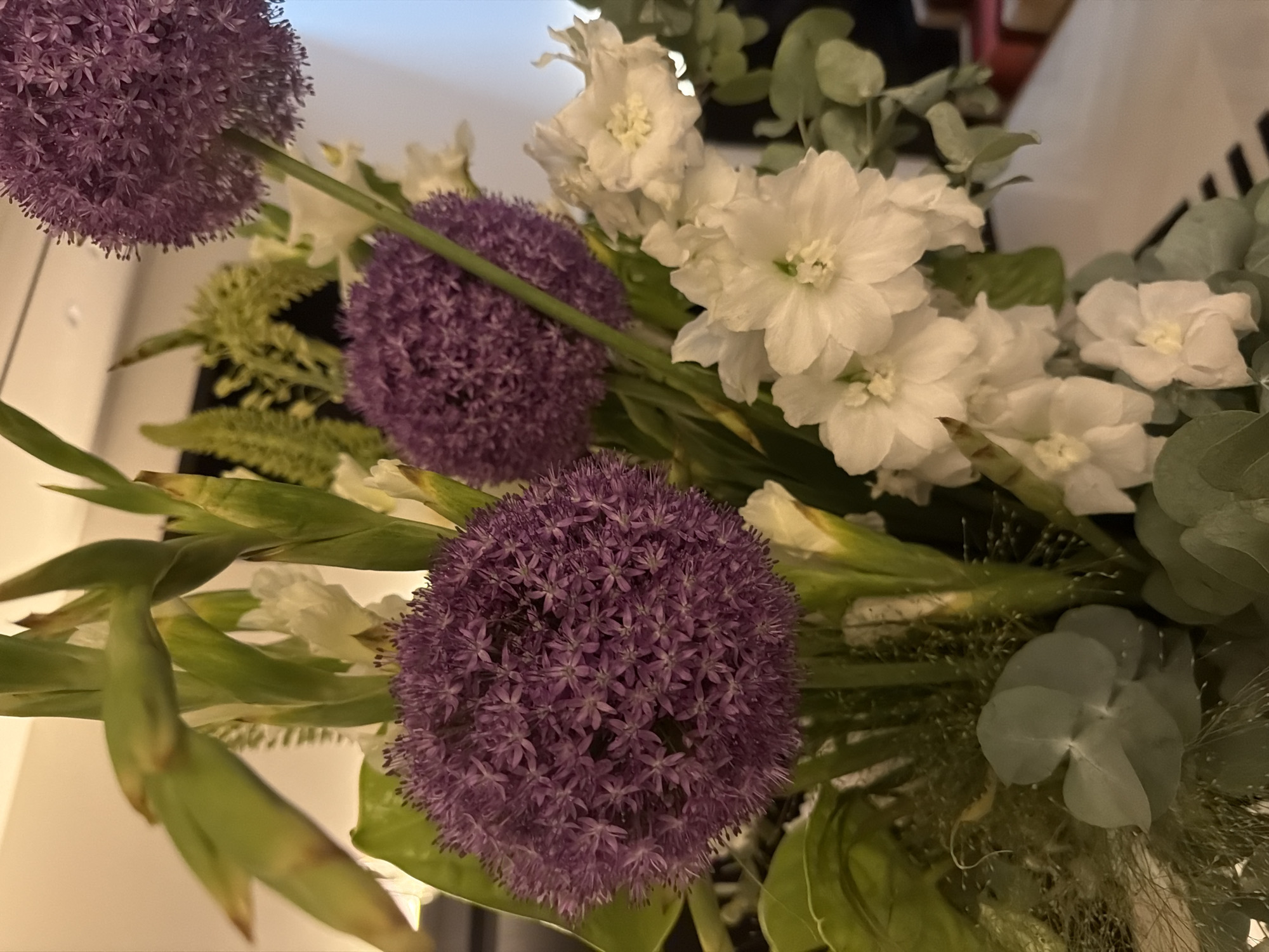 The image displays a close-up view of a floral arrangement. The arrangement features several spherical, purple allium flowers and clusters of delicate white flowers, possibly stock or delphiniums. Interspersed are green gladiolus leaves and wispy fillers. The lighting suggests an indoor setting, possibly a home or event space. There are no people or specific landmarks visible that would indicate a location within Moscow, Russia. The composition focuses solely on the flowers.