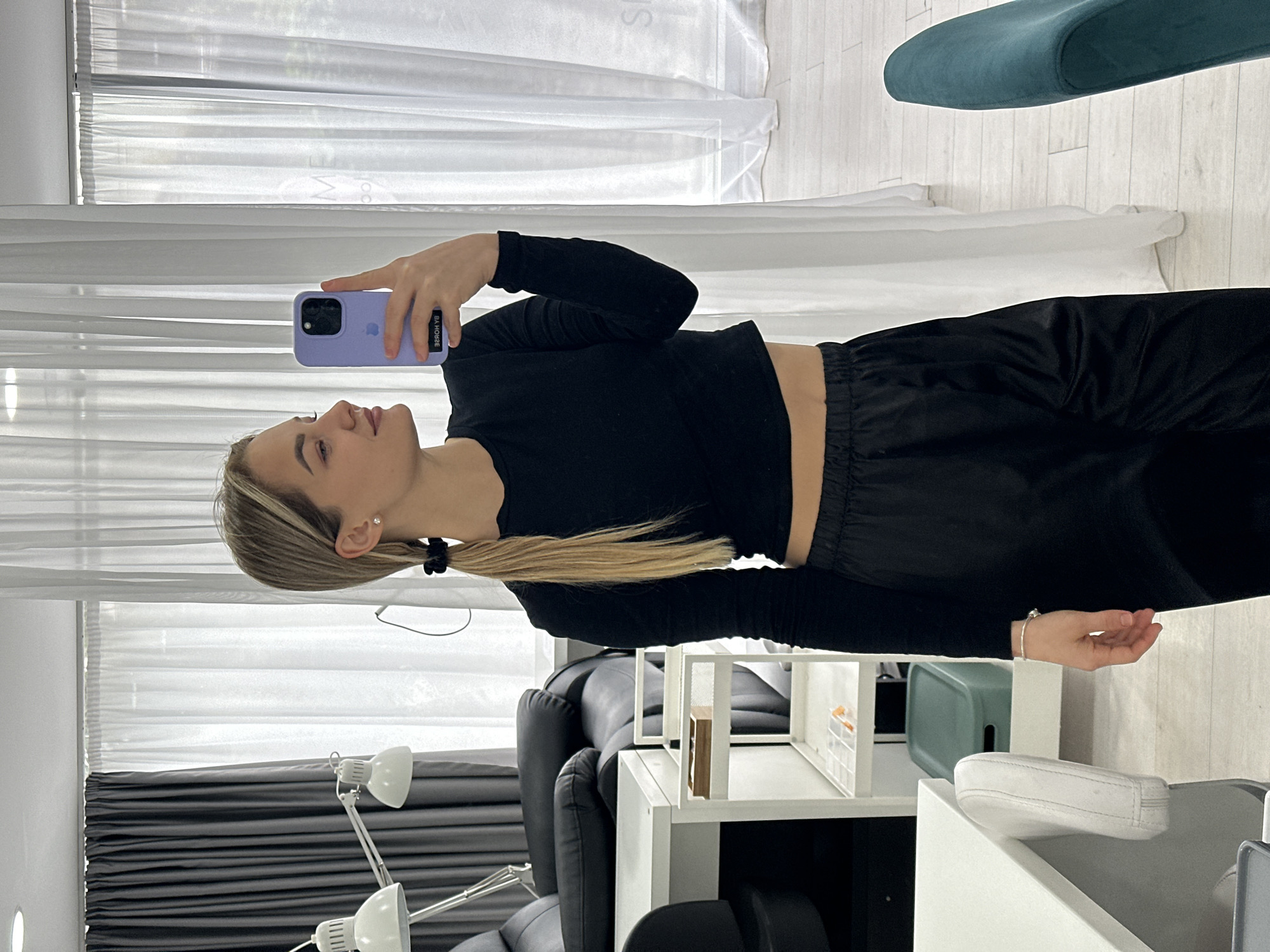 An indoor scene within a beauty salon or aesthetic clinic in Budva, Montenegro. The setting features a modern, brightly lit room with light-colored flooring and large windows covered by sheer white and dark grey curtains.
A young adult female, with blonde hair styled in two low ponytails secured with black scrunchies, is the sole individual present. She is wearing a black long-sleeved crop top, exposing her midriff, and black loose-fitting pants. She has a single diamond stud earring in her visible ear and a silver-colored bracelet on her left wrist.
The woman is taking a mirror selfie using a light lavender-cased Apple iPhone held in her right hand. Her head is turned slightly to her right, and she is looking away from the phone's lens, possibly towards her reflection or another point in the room, displaying a subtle smile.
The surrounding environment includes professional salon equipment: a black, padded reclining chair or treatment bed, two white adjustable desk lamps with slatted shades, and a white multi-tiered rolling cart holding small items, including a wooden tray on the top shelf. A partially visible white desk and a white chair with a curved backrest are to the left. A prominent teal-colored upholstered chair is positioned to the right in the background.
Reflected in the mirror where the selfie is being taken are portions of text: "ME" and "ЗЯЯОН YB" (reversed), indicating the original text "BY HOUSE." This text identifies the location as a business, specifically a beauty salon, likely "Beauty House," located in Budva, Montenegro.