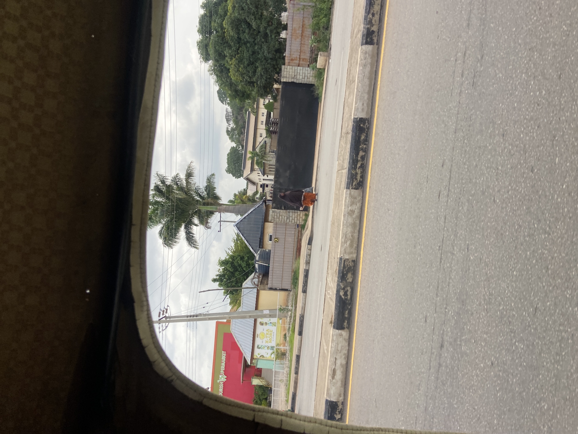 The image captures a street scene in Jos, Nigeria, viewed from within a vehicle, likely a keke-napep, indicated by the curved frame in the foreground. The foreground shows the textured interior of the vehicle and the open viewing area. The middle ground reveals a paved road with distinct black and white curb markings and a yellow line. On the sidewalk, a lone individual, appearing to be a woman wearing a dark brown hijab and an orange skirt, walks away from the viewer. Behind her, a large black gate stands closed, flanked by stone pillars and a metal fence. Adjacent to the gate is a small building with a light yellow facade and a grey metal roof, featuring a sign with green and yellow graphics. Further in the background, a bright red building with a sign reading "NOURISH SUPERMARKET" is visible. A tall palm tree stands prominently near the supermarket. The environment appears to be a commercial or mixed-use area in Jos, with residential or business structures lining the street under a cloudy sky.