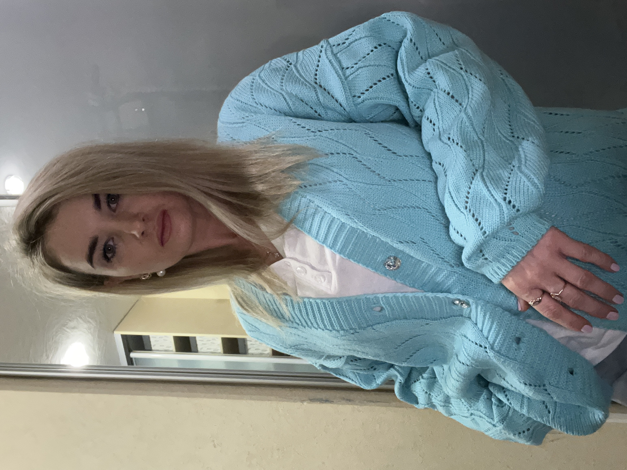A woman with light brown hair, light complexion, and wearing a blue knitted cardigan over a white collared shirt stands indoors. She has rings on her fingers and is facing the camera. The background includes a mirrored surface and a light-colored wall with a piece of furniture visible behind the mirror. There is no information in the image to determine the location in Chaikovsky, Russia.