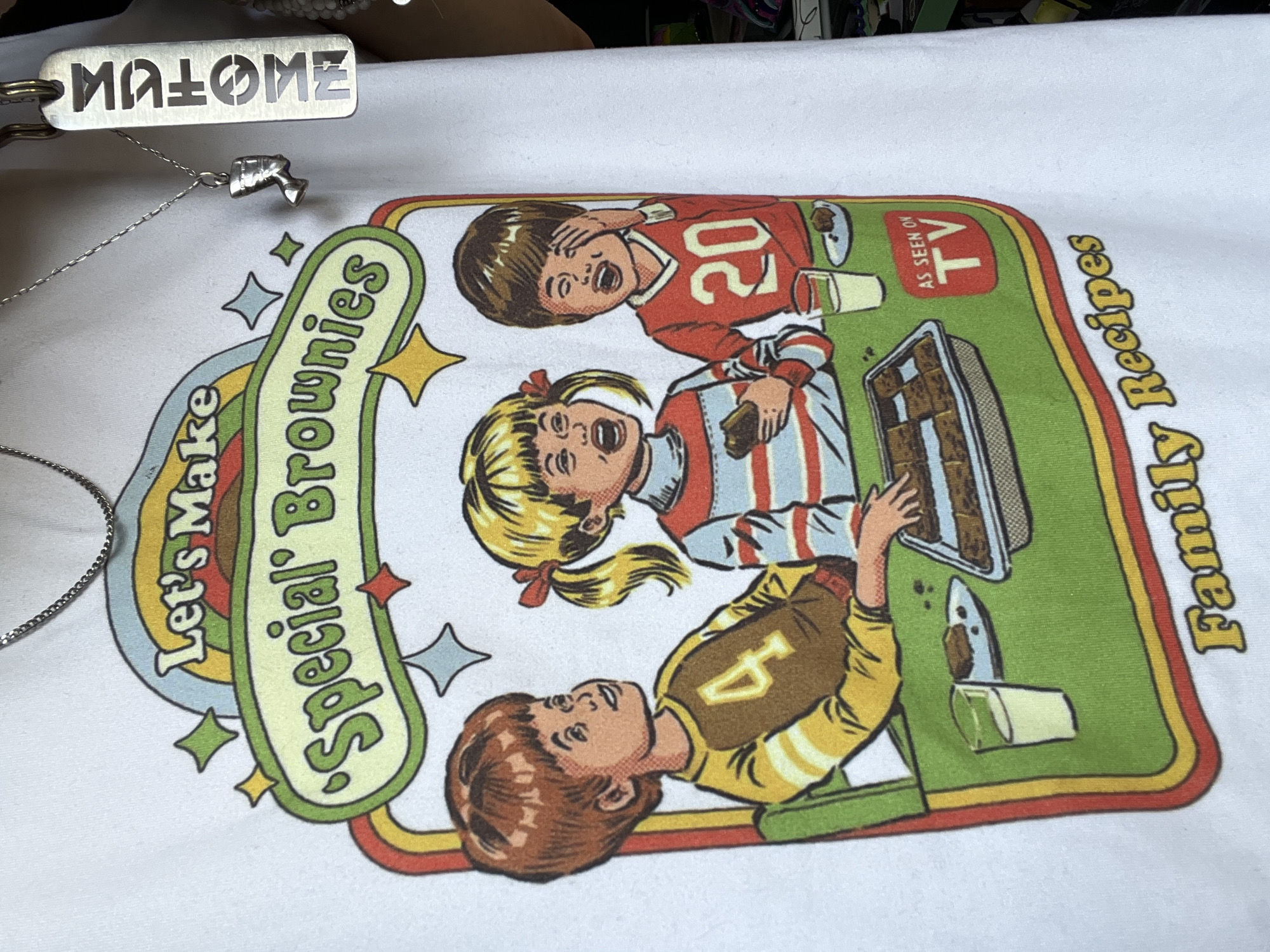 A close-up shot shows a white t-shirt with a graphic design of three children making brownies. The t-shirt is worn by a person whose neck and upper chest are visible, with a silver chain around their neck. A small pendant of a profile of a pharaoh's head hangs from the chain. Another chain with a rectangular tag is also visible. The graphic on the t-shirt features a retro-style banner that reads "Let's Make 'Special' Brownies." Below the banner, three children are depicted sitting at a table with a pan of brownies and glasses of milk. The girl in the middle holds a brownie, and the boy on the right is covering his eyes with his hand. The text "AS SEEN ON TV" is in a red box on the right side of the graphic, and "Family Recipes" is written in yellow script at the bottom. The image does not provide any information about the city of Sofia, Bulgaria, or any specific location.