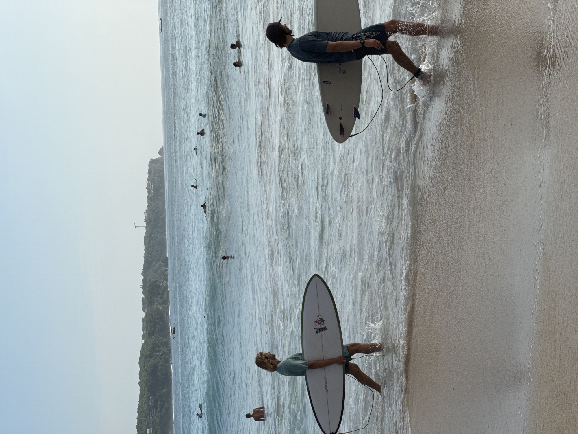 The image shows a beach scene in Kuta Selatan, Indonesia. Two surfers stand in the shallow water near the shoreline, each holding a surfboard. In the ocean, there are several people swimming or wading. Lush green foliage covers the coastline in the background under a hazy sky. The beach sand extends into the frame on the right side.