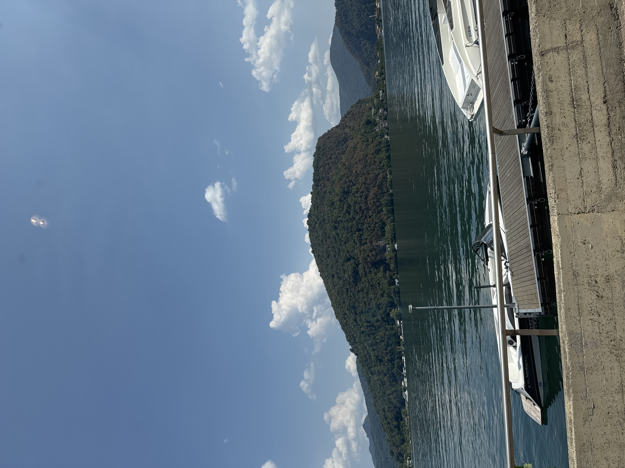 This image captures a serene lake scene with a mountainous backdrop under a clear blue sky dotted with fluffy white clouds. In the foreground, a concrete embankment runs along the bottom of the frame, with a metal railing above it. Two boats are docked at a wooden pier: a white Nautiques boat on the left, and a sleek white motorboat on the right, displaying the registration number "CSG 6142". The lake's surface is calm and reflects the surrounding landscape. The forested mountains rise majestically from the water's edge, with a scattering of buildings visible along the shore. The overall impression is one of tranquility and natural beauty, characteristic of the lakeside setting in Lavena Ponte Tresa, Italy.