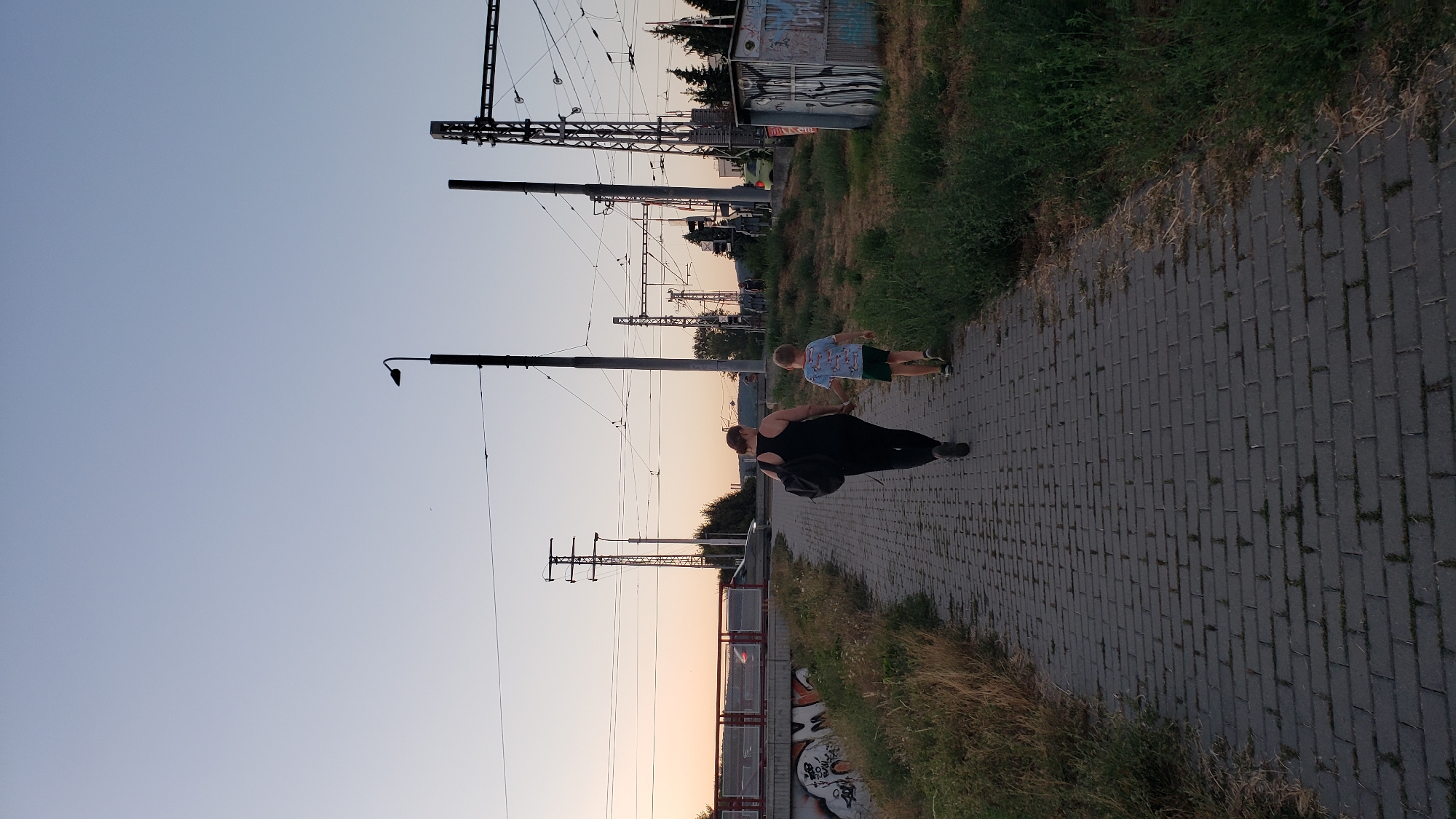 In Prešov, Slovakia, a woman and a young boy walk hand-in-hand on a paved sidewalk beside a grassy area. The woman, seen from behind, wears a black tank top and dark pants, carrying a black backpack. The boy, also viewed from behind, is dressed in a blue t-shirt with a pattern and green shorts. They are walking away from the viewer towards a set of railway tracks with overhead electrical lines. In the background, a bridge with a car crossing and graffiti-covered structures are visible. The sky is a pale blue, transitioning to a warm orange hue near the horizon, suggesting either sunrise or sunset. The scene is captured outdoors, with visible vegetation and industrial-style infrastructure.