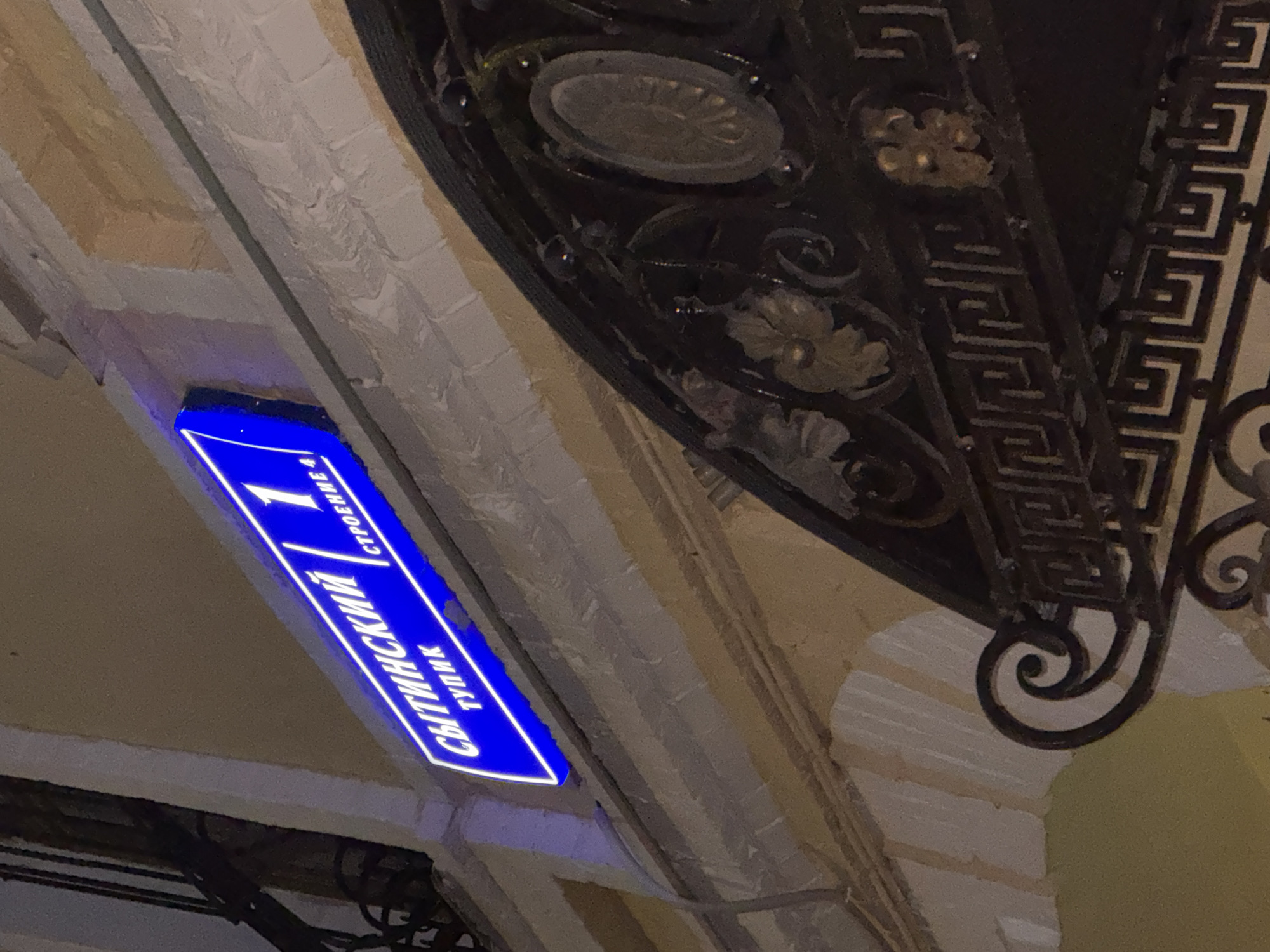 The image shows a blue street sign with white lettering, indicating "СЫТИНСКИЙ 1 ТУПИК СТРОЕНИЕ 4" which translates to "Sytinsky 1 Dead End Building 4". The sign is illuminated with neon blue light. It is mounted on a textured, light-colored building facade. Below the sign, an ornate, dark metal structure, possibly an awning or gate, is visible, featuring decorative scrollwork and geometric patterns. The scene appears to be outdoors at night, given the artificial lighting and the dark surroundings. The location is Moscow, Russia.