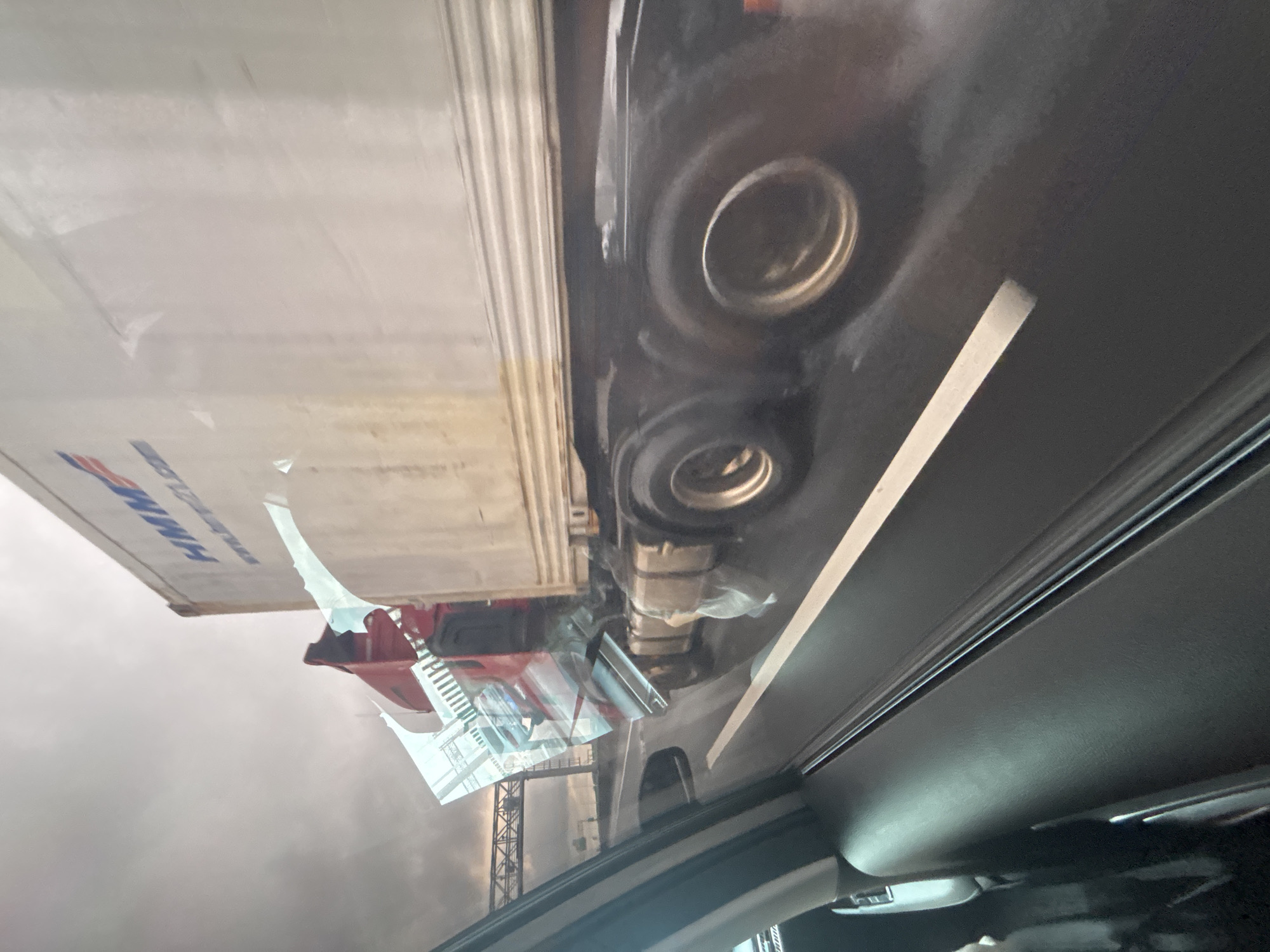 The image shows a view from inside a vehicle looking out at a damaged truck on a wet road. The truck appears to be a semi-trailer truck with its trailer severely damaged and tilted. The front of the truck, the red cab, is visible and also appears to have sustained damage, with glass broken and some deformation. Reflections on the truck's trailer suggest it is a light-colored, ribbed metal container. The road surface is wet, indicating recent rain or spray from other vehicles. In the background, an overpass or bridge structure is visible under a cloudy sky. The scene appears to be on a highway or major road, likely in an urban or suburban area given the presence of the overpass. The photograph captures the aftermath of a traffic incident, with the damaged truck a central focus. There are no people visible in the image. The location is identified as Baiyun District, China.