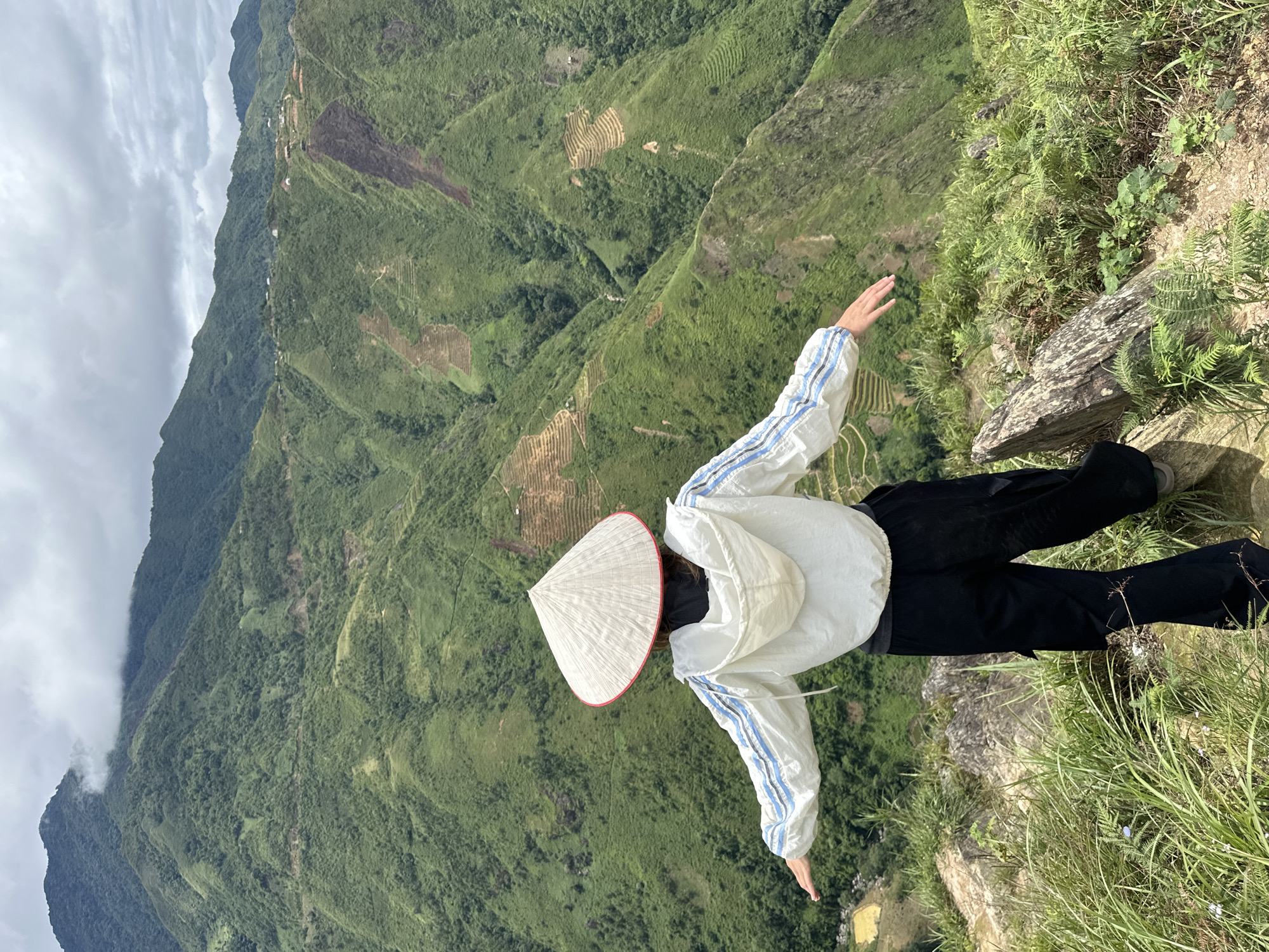 A person wearing a conical hat and a white jacket with blue stripes stands on a rocky outcrop, facing away from the camera, with arms outstretched. The person is dressed in black pants. The background is a vast, lush green mountainous landscape with terraced fields and a cloudy sky. The scene appears to be in Ta Xua, Vietnam, a location known for its scenic mountainous terrain.