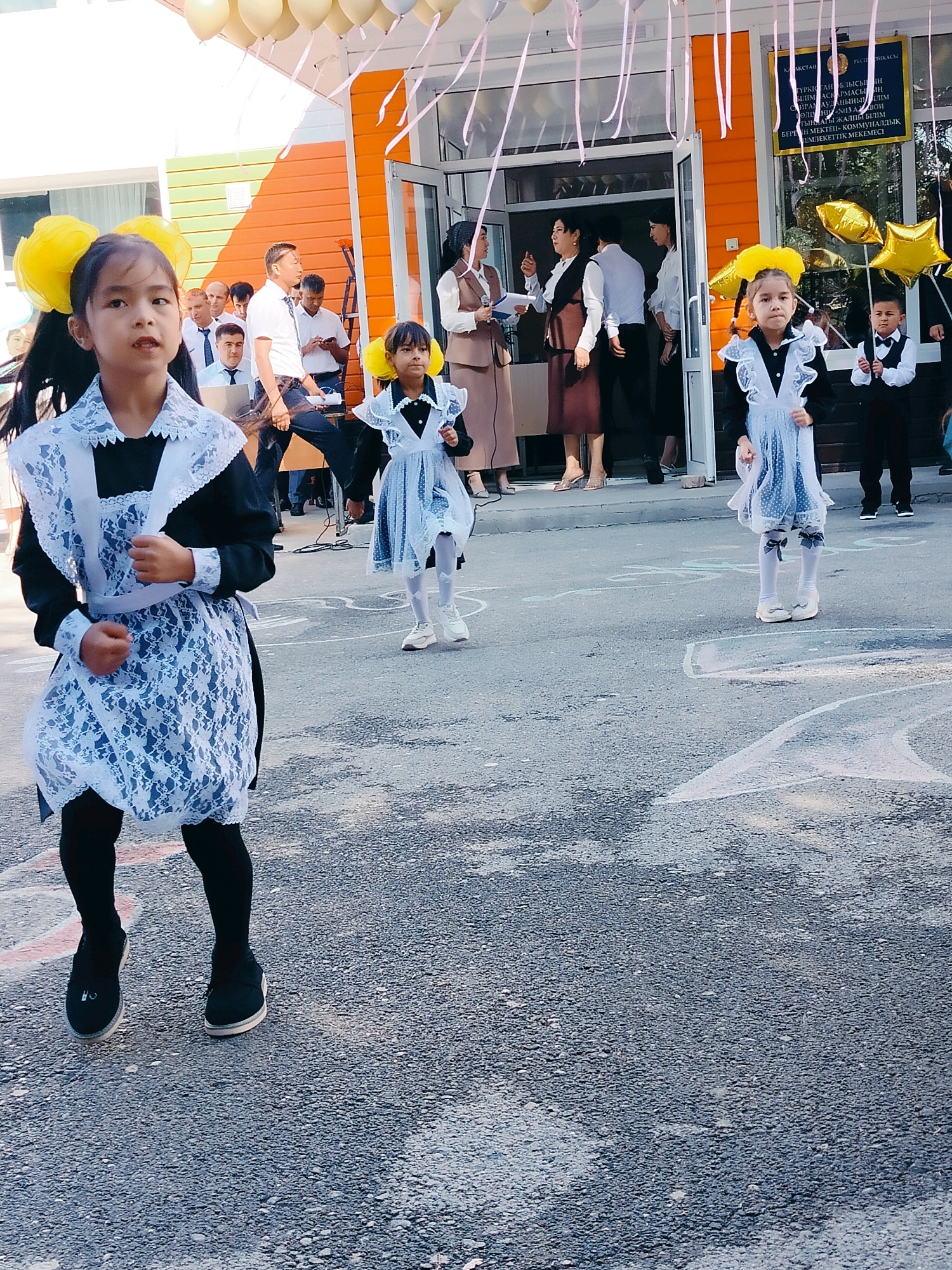 In the city of Kölkent, Kazakhstan, a group of schoolchildren are gathered outside a building, likely for a school event. Two young girls in the foreground are dressed in traditional school uniforms, featuring black dresses with white lace collars and aprons, and yellow hair accessories. They appear to be in motion, possibly dancing or performing. Behind them, other students and adults are visible, some holding balloons. The building in the background has an orange facade and an entrance with a sign in Kazakh. The ground is paved and appears to have chalk drawings on it, suggesting a festive or celebratory atmosphere.