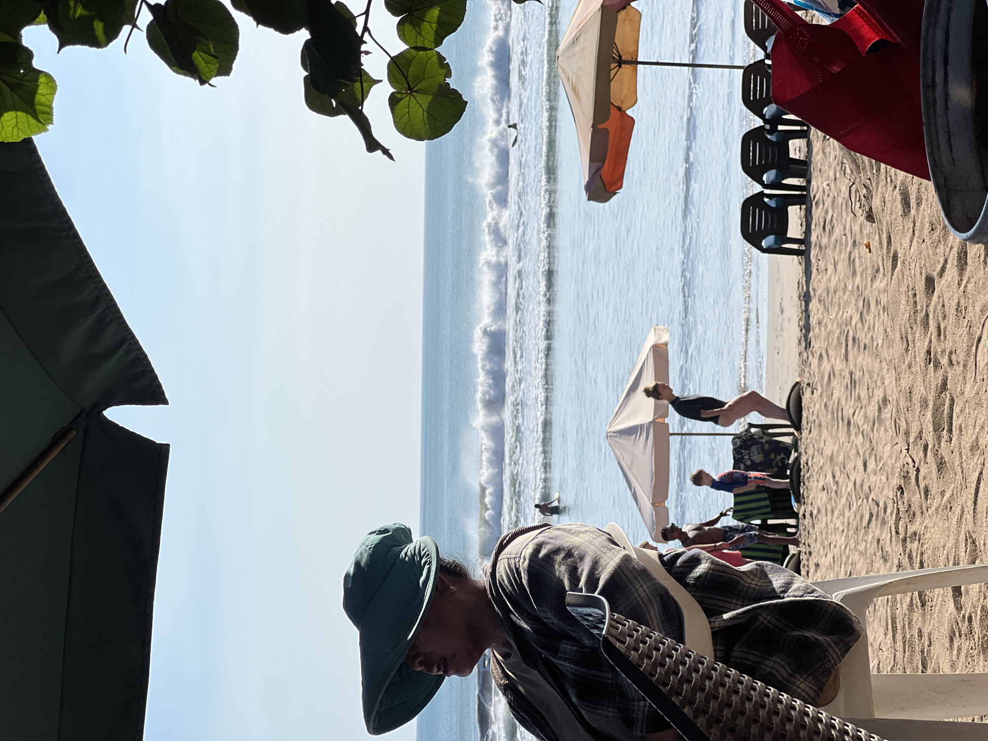 This image depicts a beach scene in Kuta, Indonesia. In the foreground, a person wearing a large green hat and a plaid shirt is seated in a white chair, facing left and looking out towards the ocean. Their body is partially obscured by the chair and their attire. Behind them, the sandy beach stretches towards the water. Several beach chairs are arranged on the sand, some unoccupied and others with people nearby.
In the middle ground, a group of individuals are visible. One person, a woman in dark attire, stands near a beach umbrella. A young boy in a blue and red outfit is also present. Further out in the ocean, waves are breaking, and a lone surfer is visible riding a wave. The ocean extends to the horizon, meeting a clear blue sky. The scene is framed by the dark canopy of an umbrella in the upper left and lush green leaves of a tree in the upper right, suggesting a shaded area overlooking the beach. The overall atmosphere is one of a relaxed beach day.