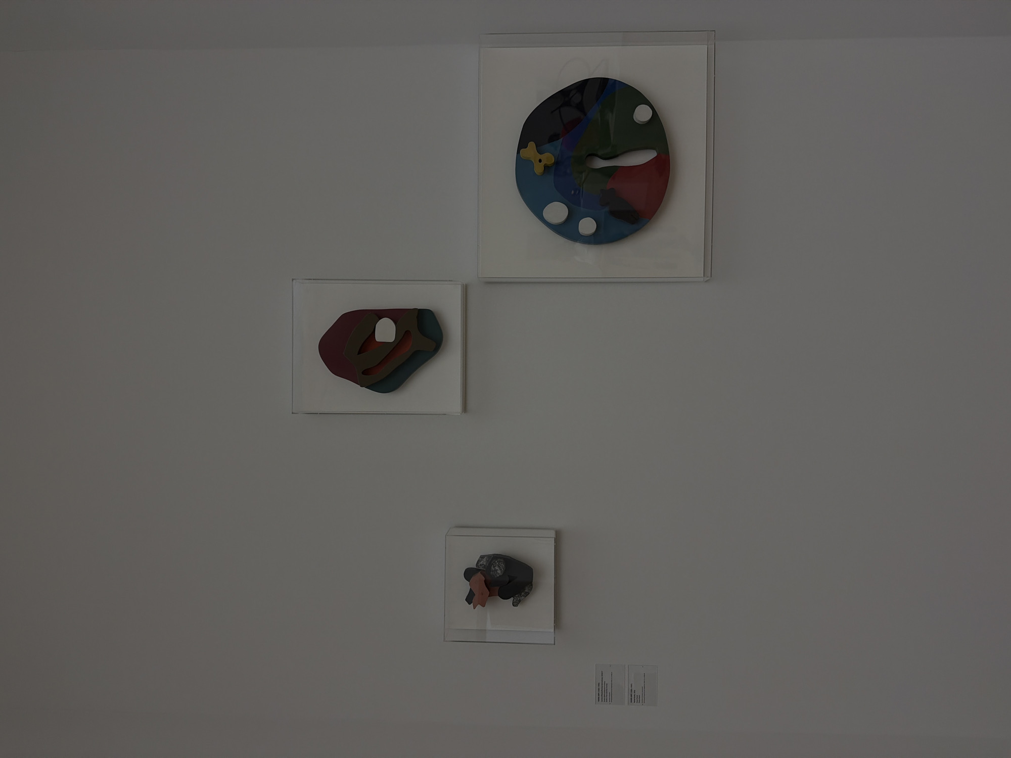 Three framed artworks are displayed on a plain white wall, typical of an art gallery setting. The art pieces are abstract and colorful. On the left, a small, square frame contains a dark gray and pink sculptural piece. Below this artwork, two white placards are mounted on the wall, likely providing information about the art. In the center, a rectangular frame holds another abstract artwork with layered shapes in shades of red, green, blue, and brown, with a white oval shape in the middle. To the right, a larger square frame displays a circular artwork composed of various colored segments, including blue, green, and red, with white circles and a yellow abstract shape near the top. The overall impression is one of a clean, minimalist exhibition space. This scene is located in the Fondation Beyeler museum in Riehen, a municipality of Basel, Switzerland.