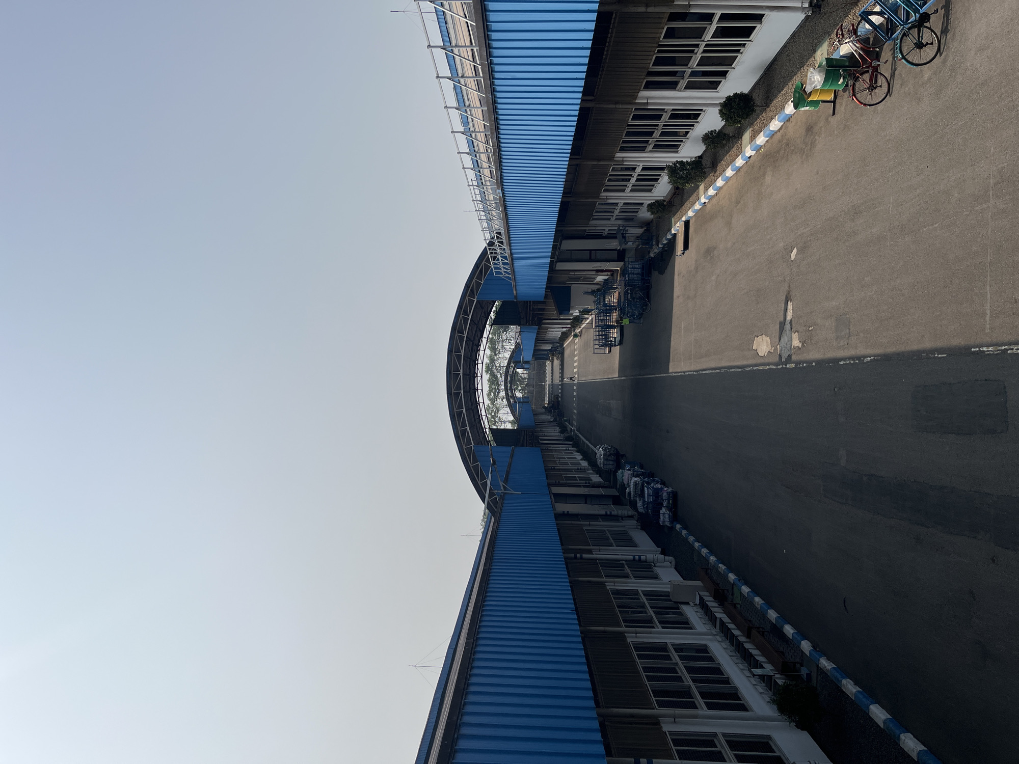 This is a forensic, matter-of-fact description of the image, taken in the city of Pelang, Indonesia.
The image depicts an outdoor industrial or warehouse area. A wide asphalt road runs through the center, flanked by long, low-lying buildings with blue corrugated metal roofs and white siding. The buildings have numerous windows. Along the sides of the road, there are various objects including several trolleys, some appearing to be loaded with goods, and a few bicycles. Two green trash bins are also visible. The sky is clear and pale. There are a few small trees and bushes planted along the edges of the buildings. The overall scene suggests a functional, operational facility under daylight conditions.